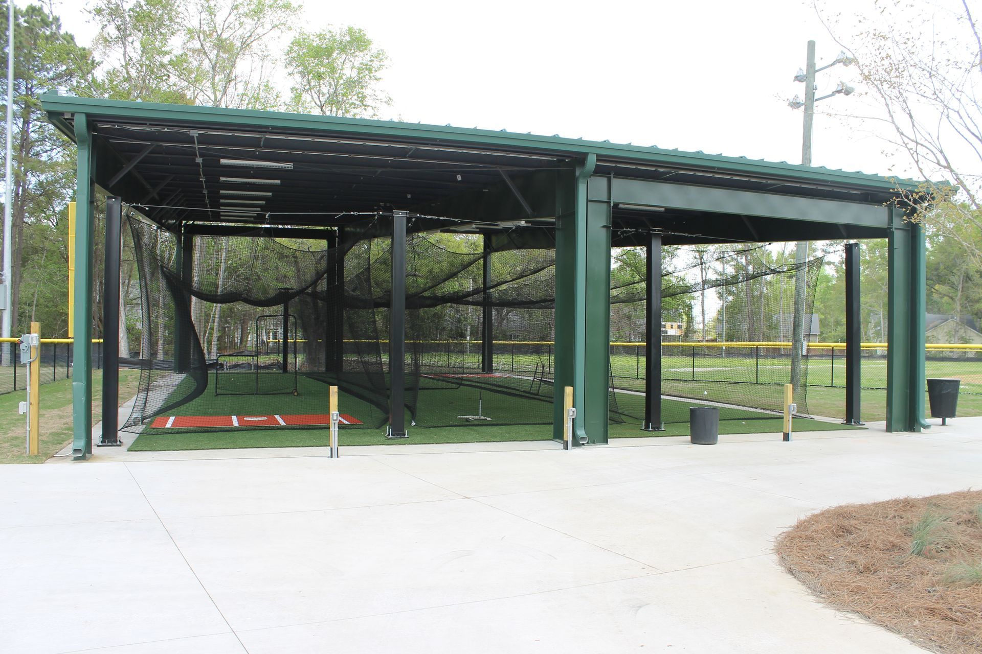 Baseball batting cage shelter with green metal roof and supports, black netting, and artificial turf.