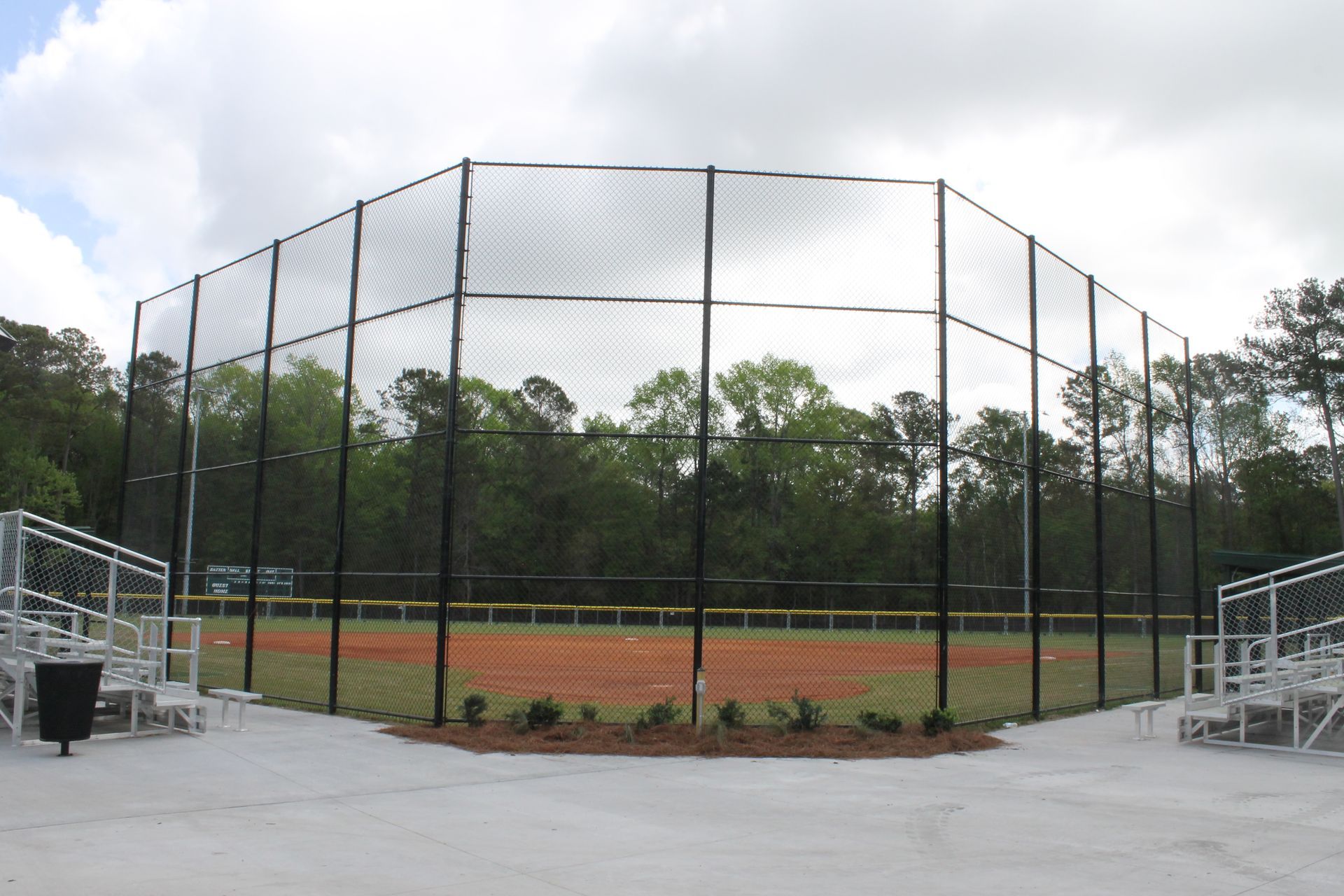 Baseball field with black safety netting, bleachers, and red dirt infield. Trees in background.