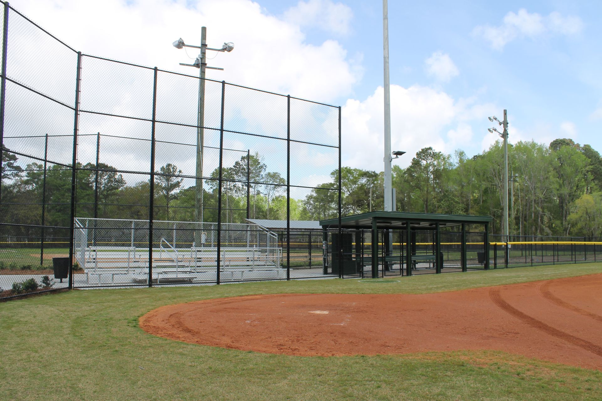 Baseball field with a chain-link fence, dugout, and netting. Brown dirt field, green grass, and trees in the background.