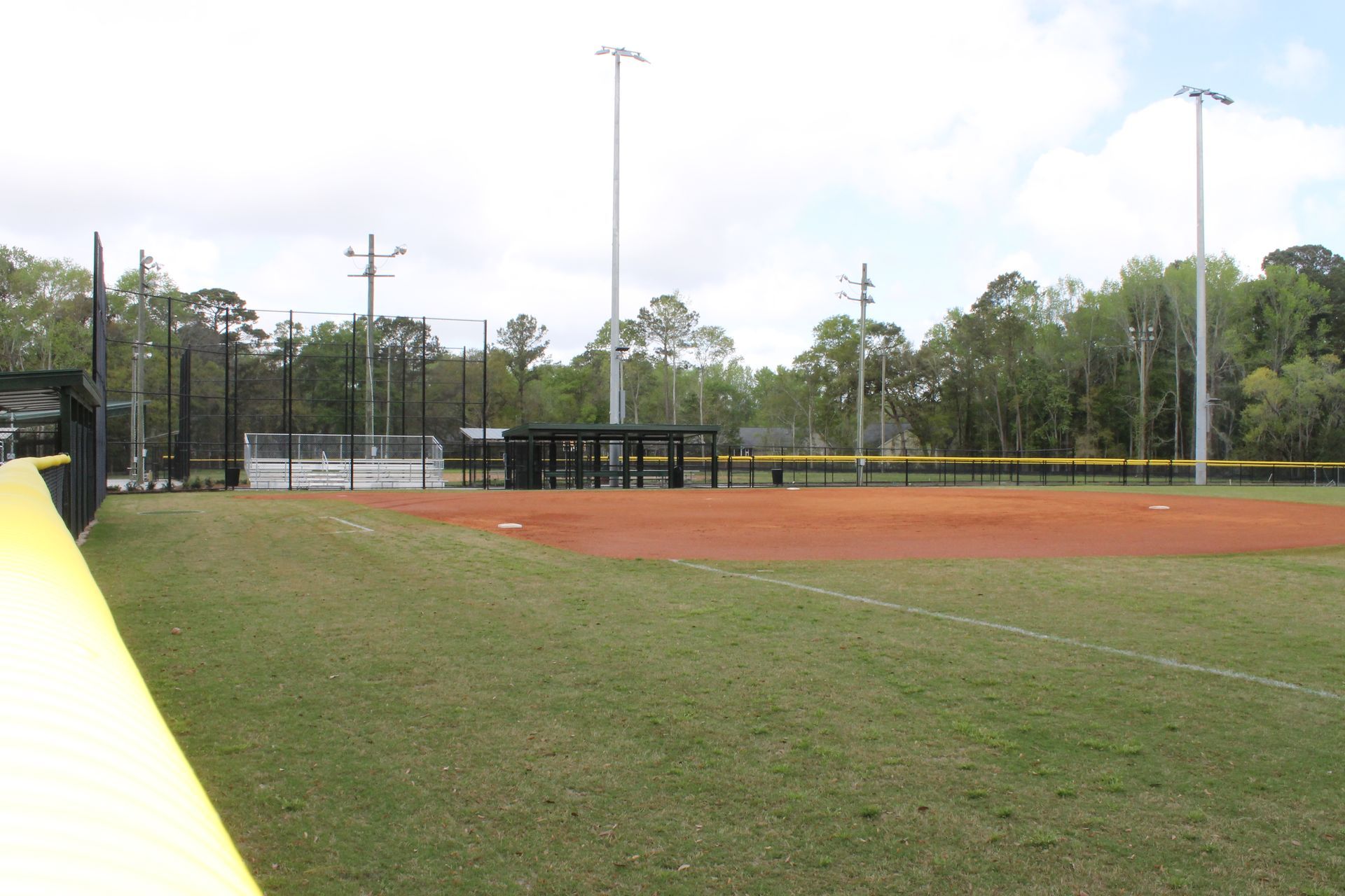 A baseball field with green grass, red dirt, and black fencing in front of trees under a cloudy sky.