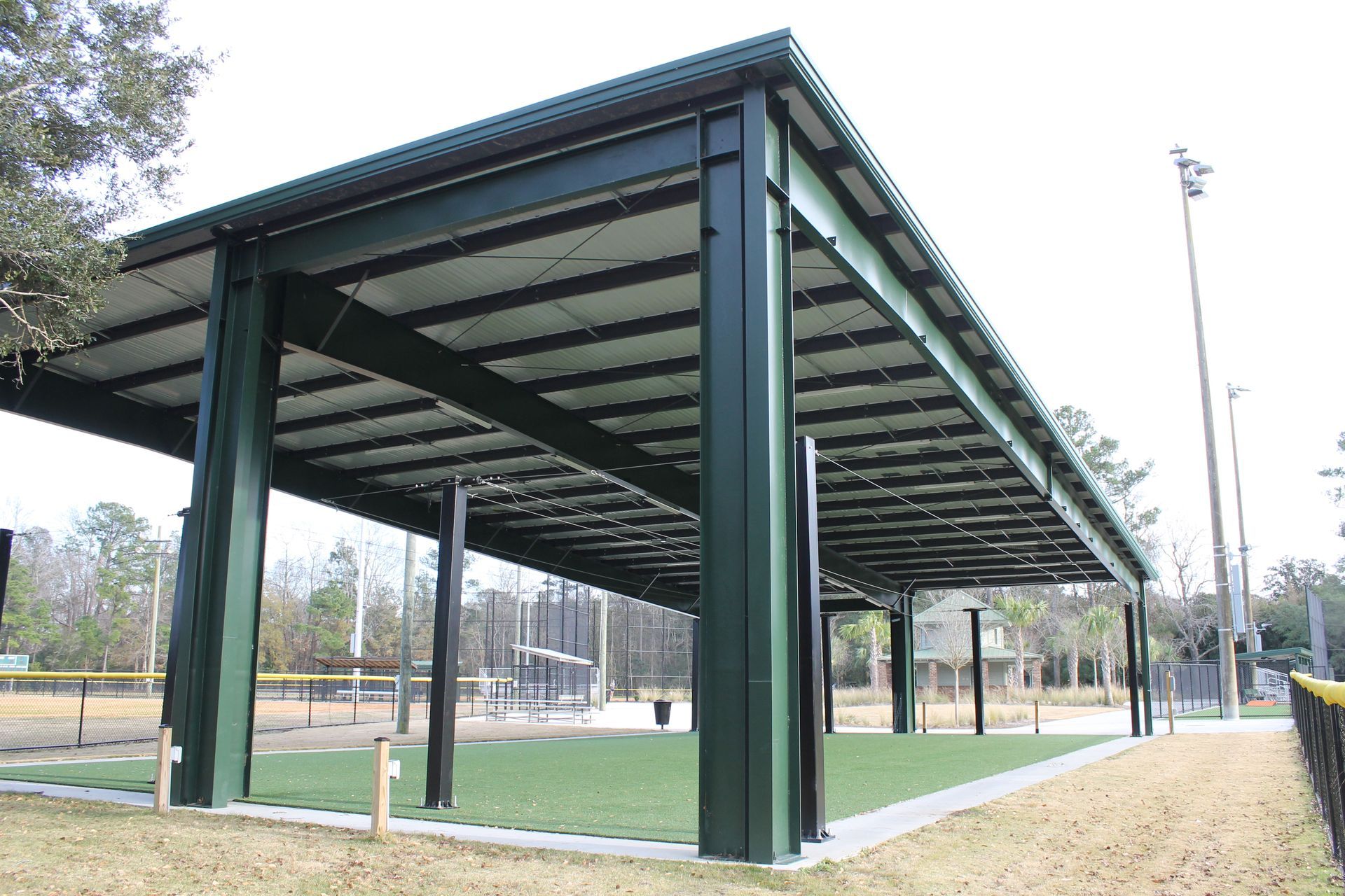 Green covered shelter at a baseball field with a grassy area and the field in the background.