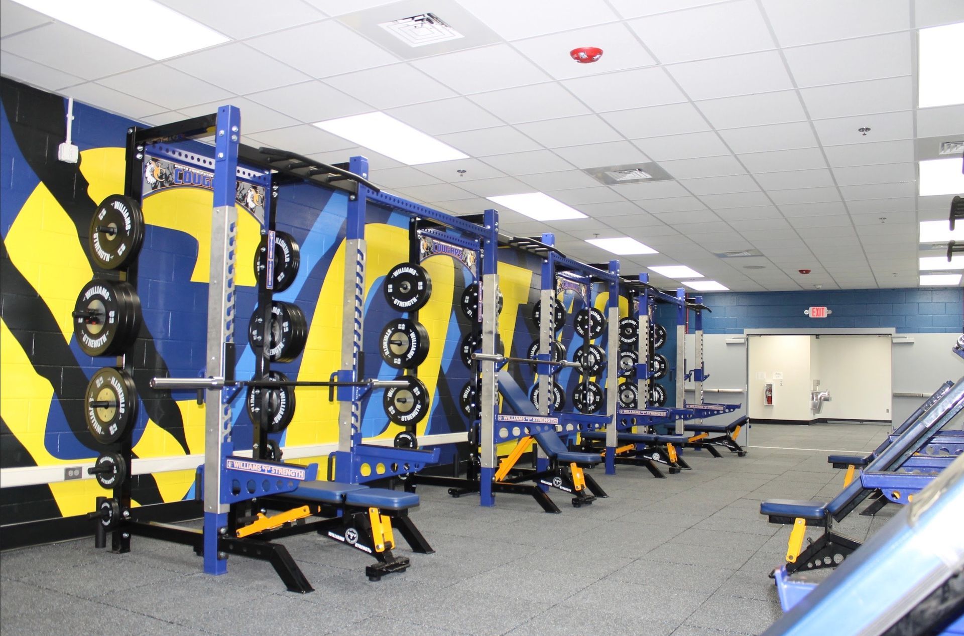 Weight room with blue and yellow weight racks, benches, and weights.