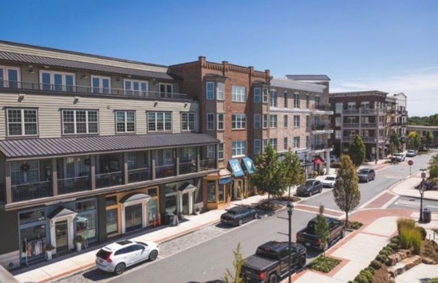 Buildings with shops and apartments line a sunny street with parked cars and trees.