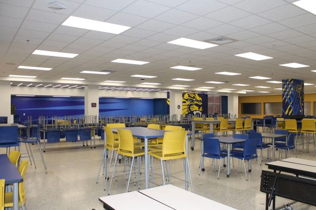 Empty school cafeteria with yellow and blue tables and chairs, white ceiling, and blue wall art.