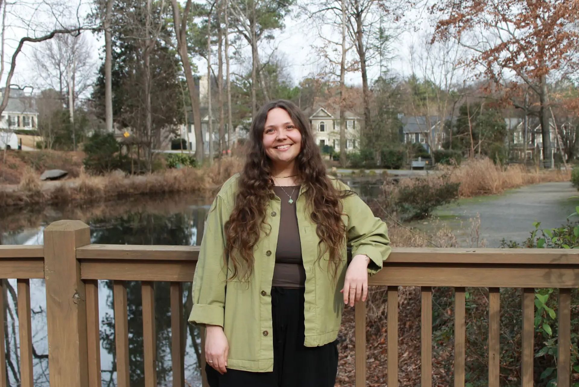 Woman with long brown hair, green jacket, standing on a wooden deck overlooking a pond and trees.