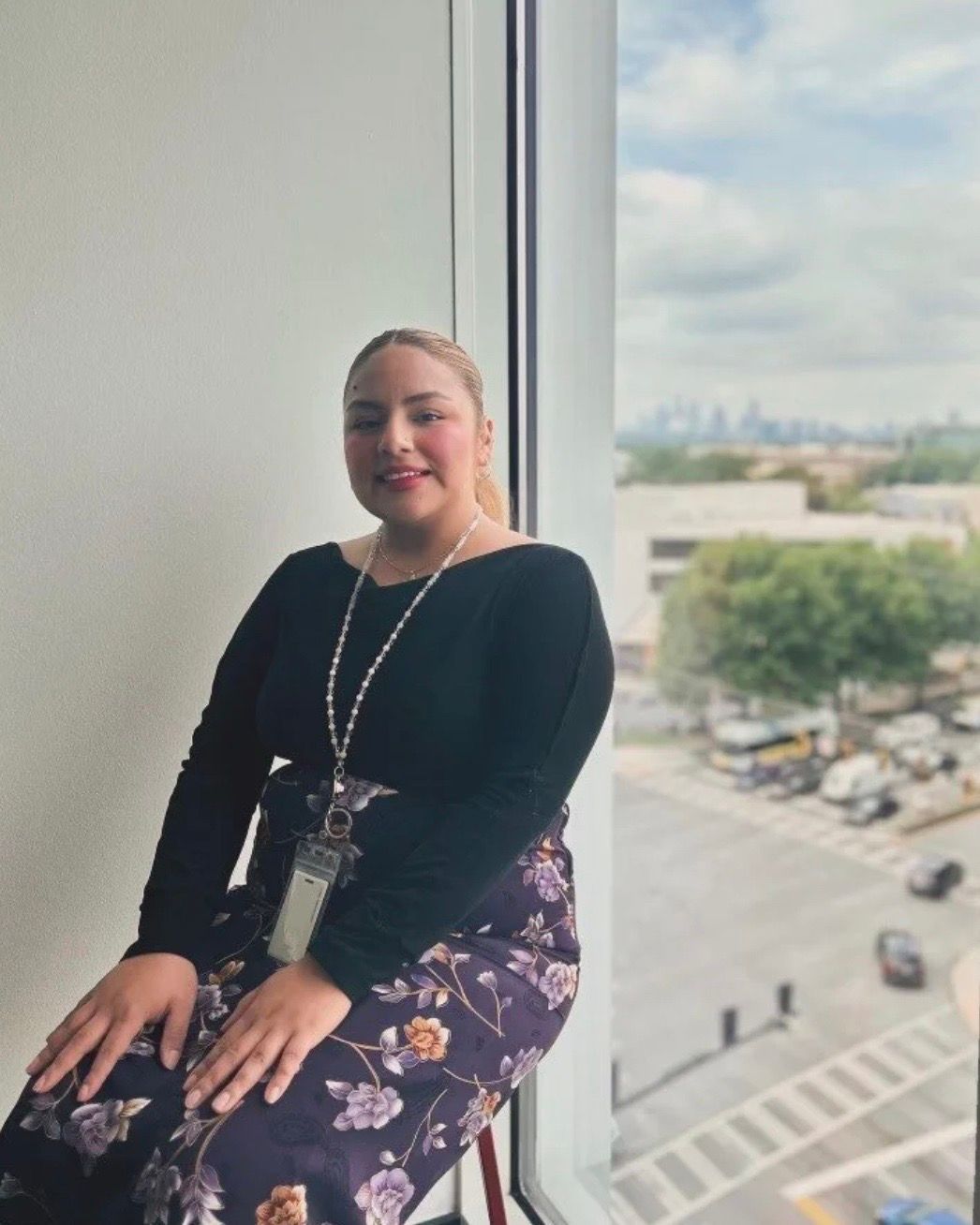 Woman in black top and floral pants sits near a window, city view in the background.