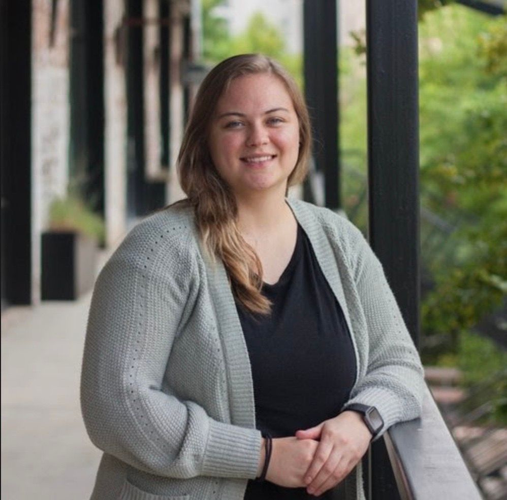 Woman in light gray cardigan smiles outdoors, leaning on a railing.