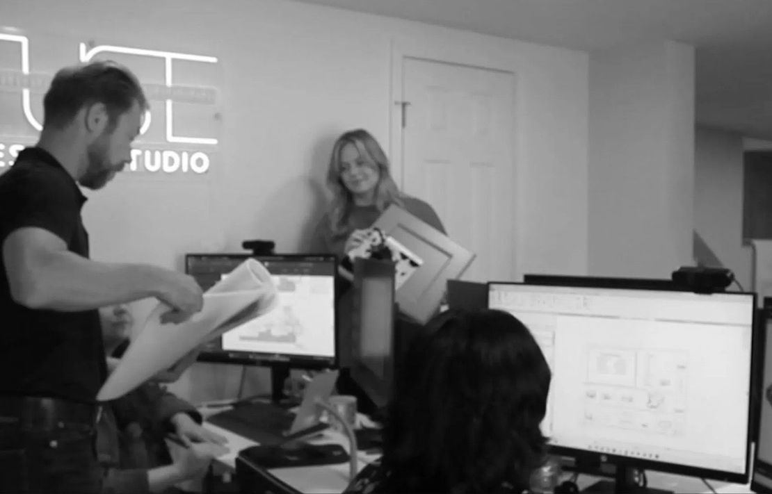 People in an office reviewing documents and computer screens. Man holding papers, woman smiling with a framed object.
