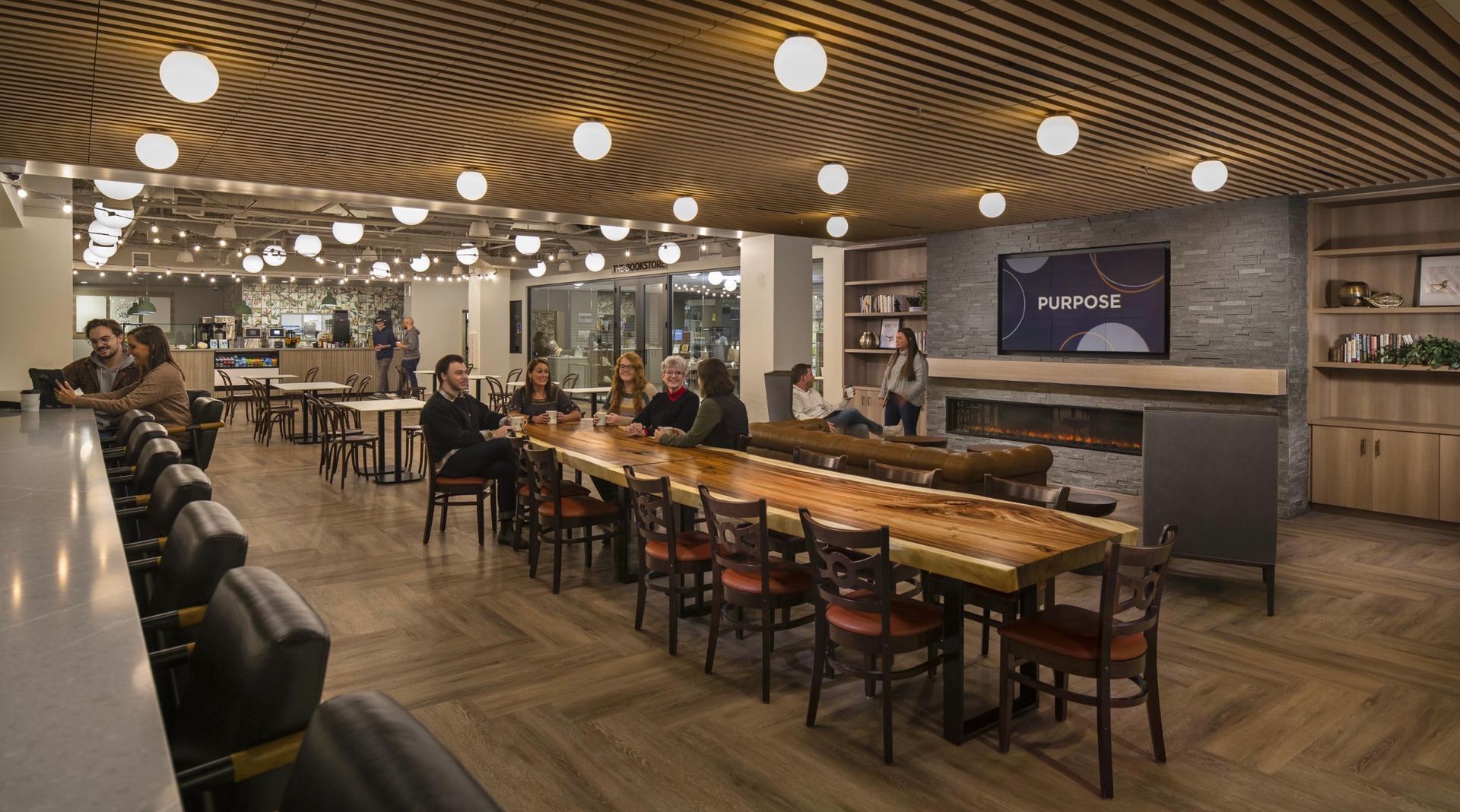 Lounge area with bar, tables, fireplace, and large screen TV. People are seated and interacting in the space.