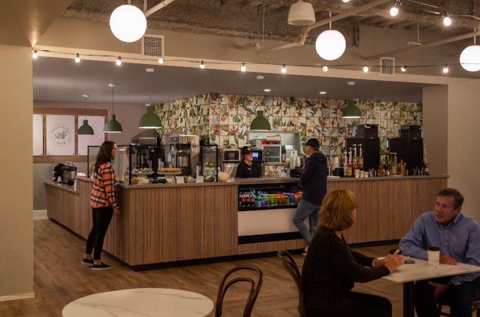 Coffee shop interior with customers and staff. Bar with drinks, food, and cashier. Wooden tables, chairs, and overhead lights.