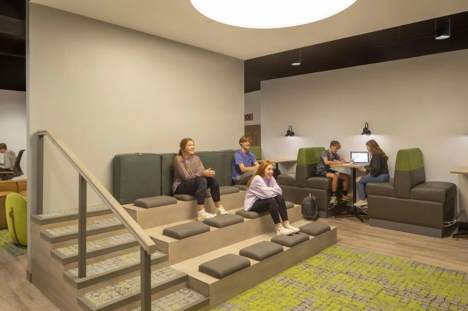 Students sitting in a modern lounge area. Gray and green seating on steps and in booths, with a green carpet.