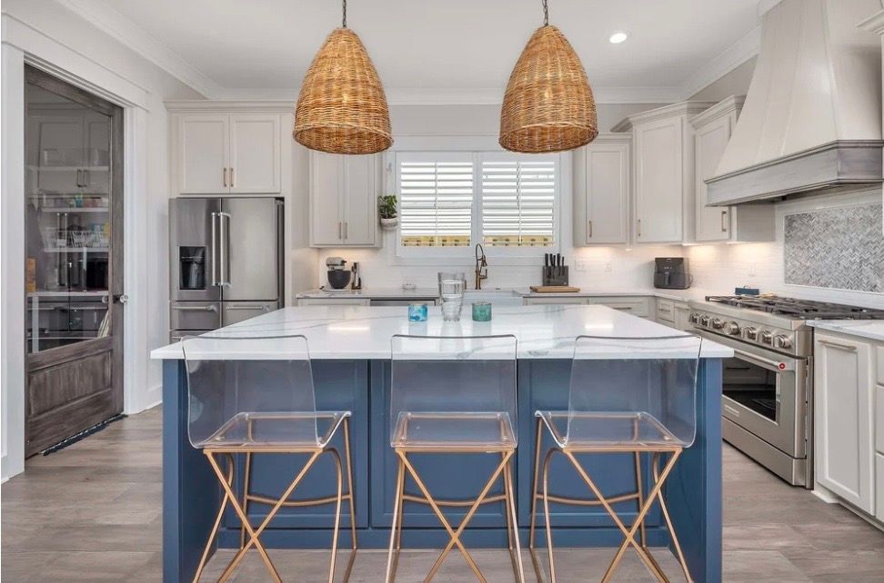 Kitchen with blue island, white cabinets, marble countertop, rattan pendant lights, and clear bar stools.