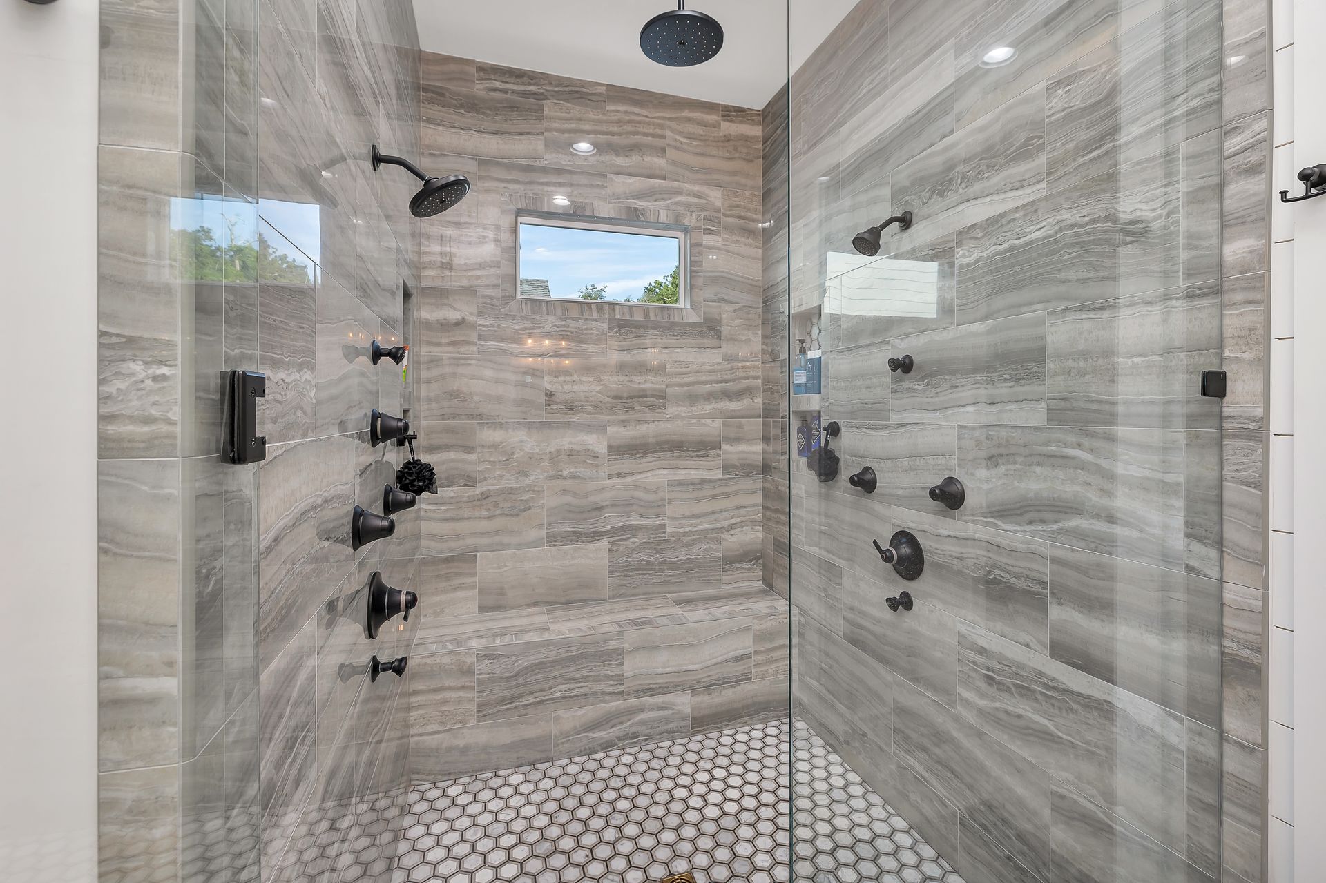Spacious modern shower with gray tile walls, pebble floor, multiple black showerheads, and glass doors.