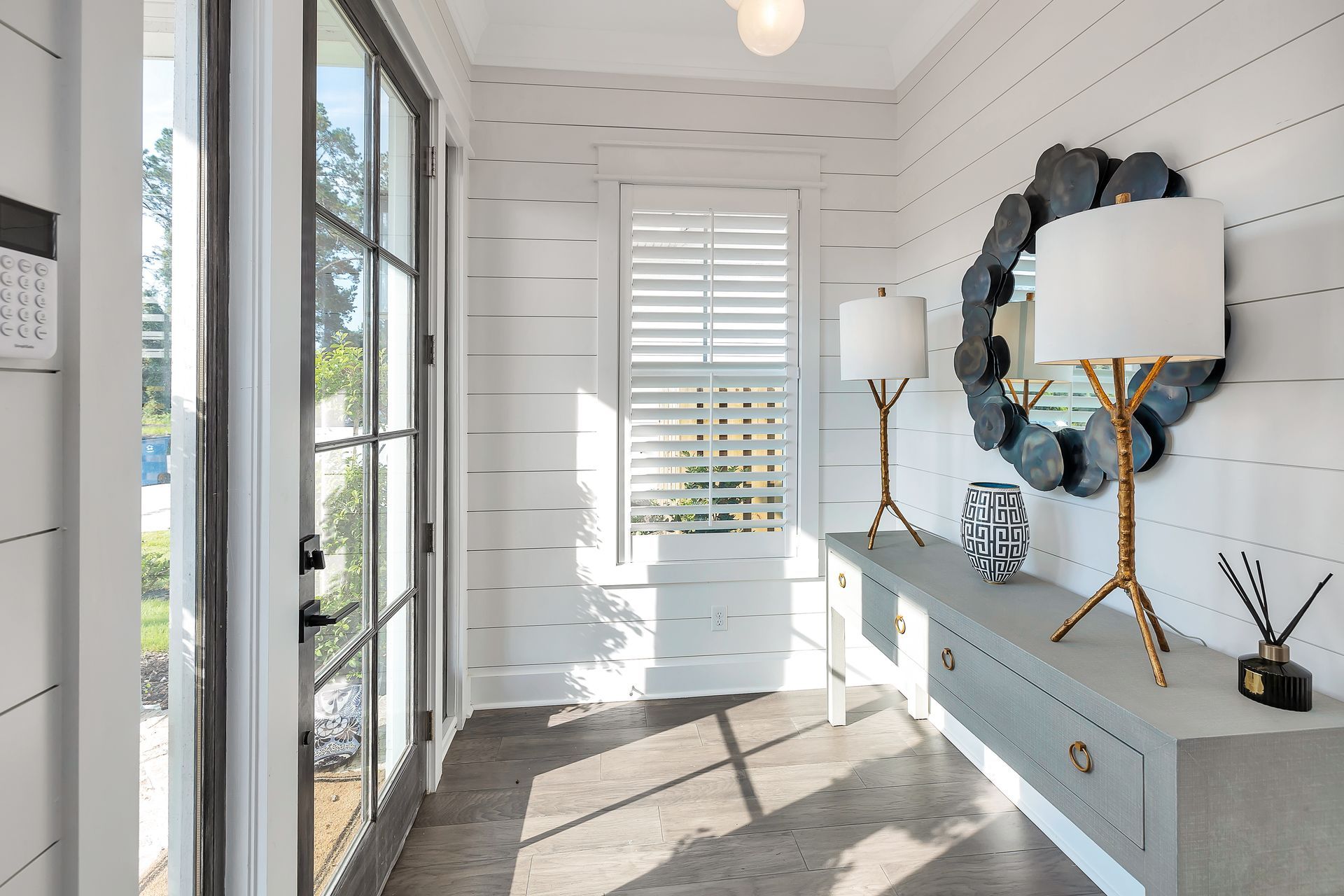 Hallway with gray console table, large mirror, lamps, and white shiplap walls. Sunlight streams in.