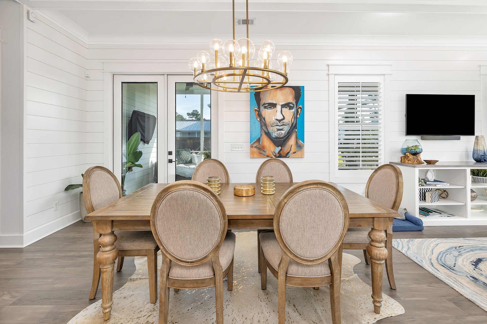 Dining room with light wood table, upholstered chairs, artwork, and chandelier.