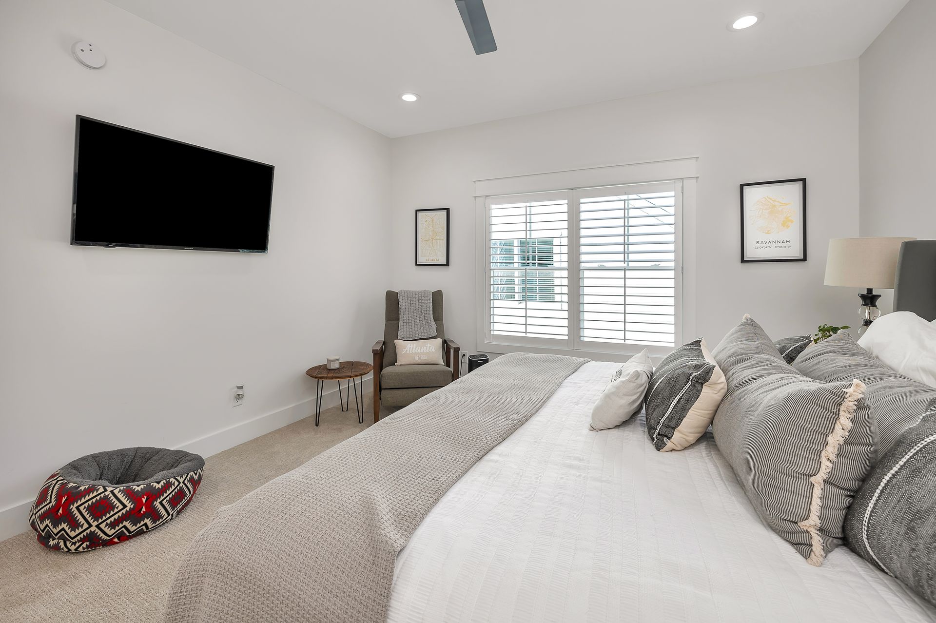 Bedroom with a bed, TV, chair, and a dog bed. White walls, window with shutters, and neutral colors.