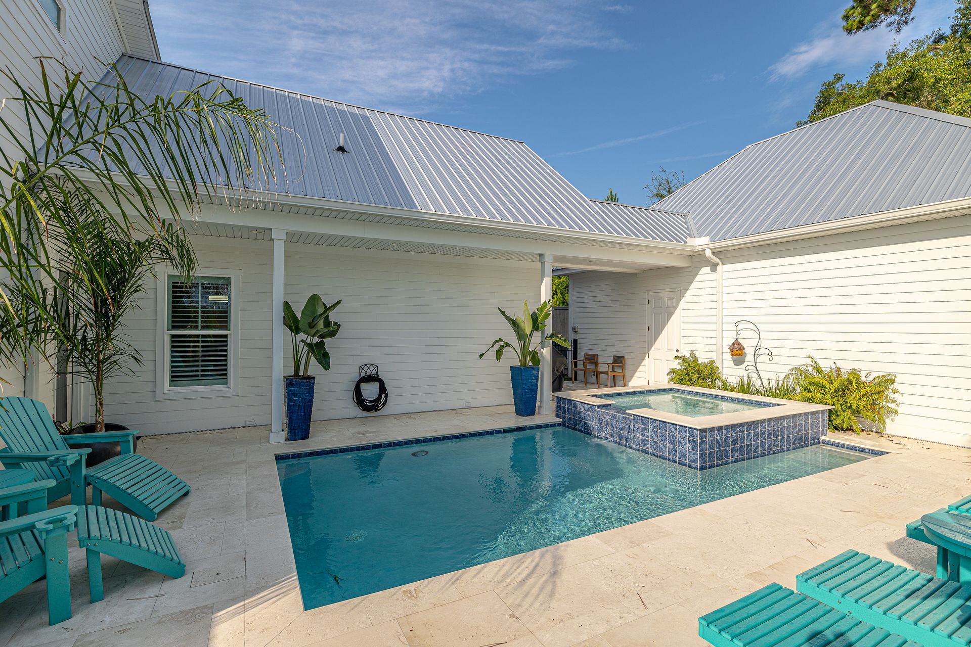 Courtyard with a pool, hot tub, white walls, and blue chairs under a bright sky.