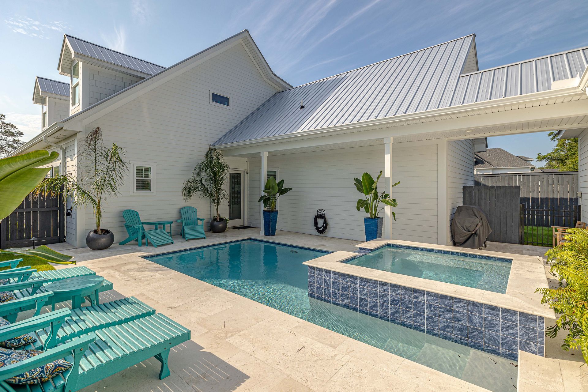 Backyard pool and spa with white house, turquoise chairs, and blue accents under a blue sky.
