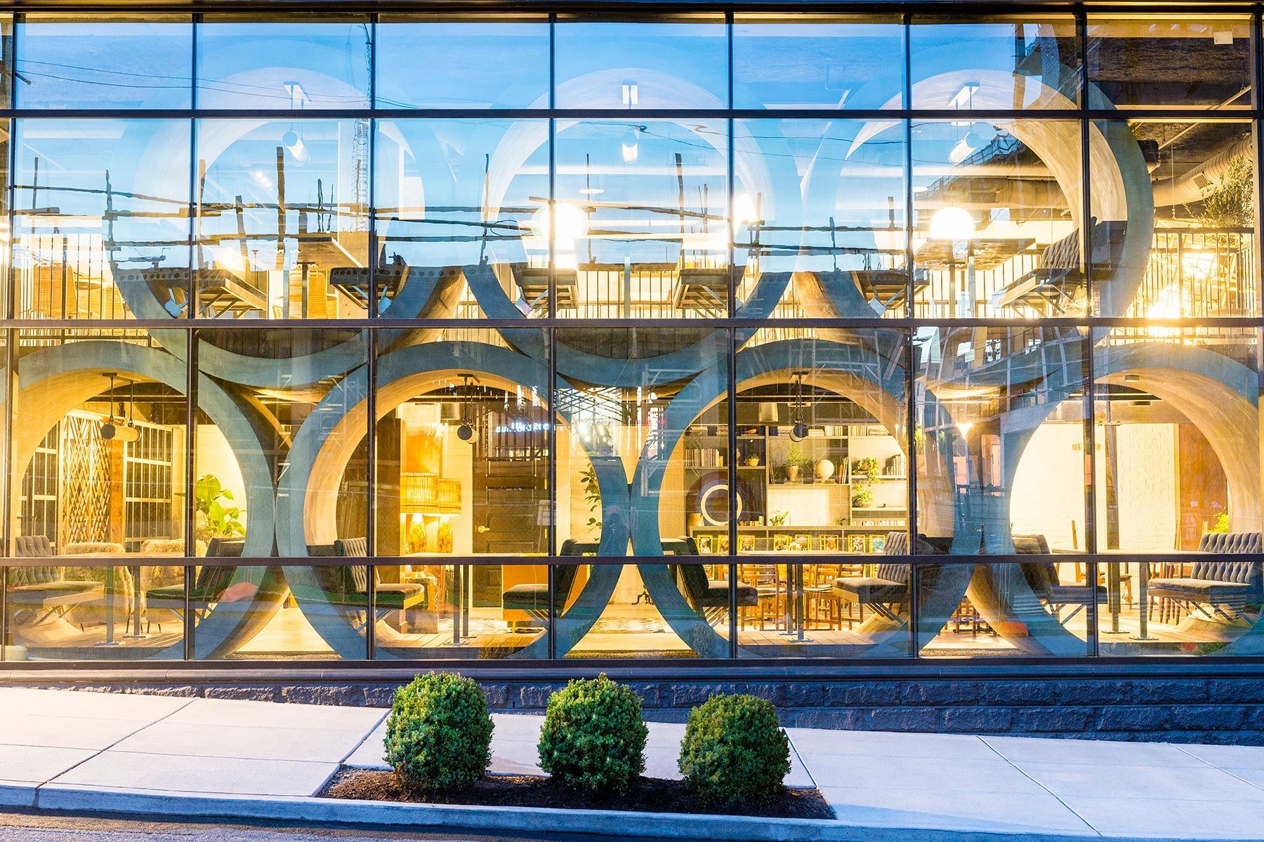 Glass facade of a building with circular design elements; interior lit, showing tables and lamps; green shrubs in front.