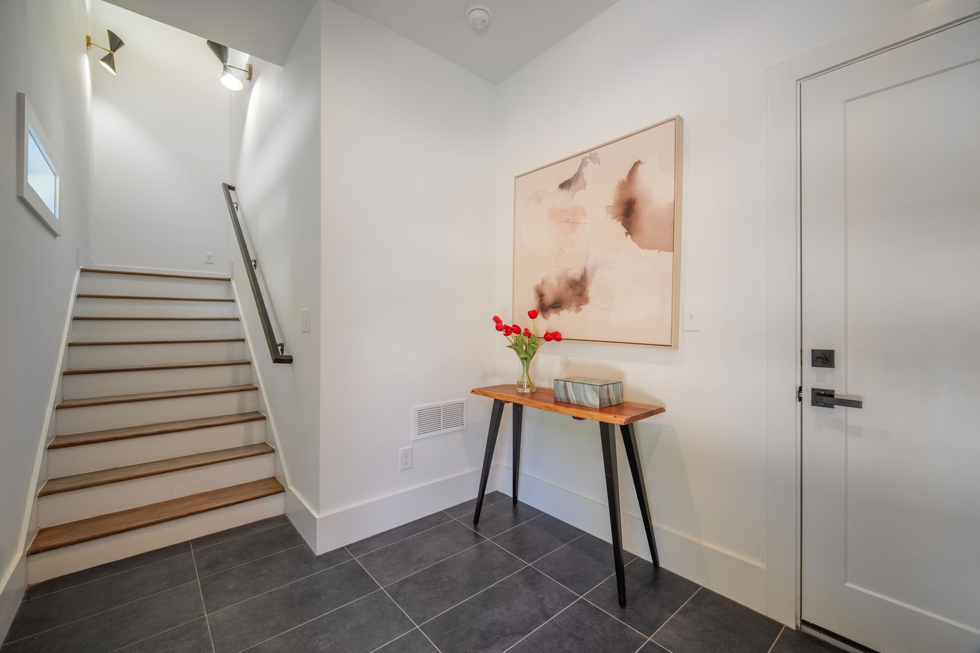 Entryway with stairs, art, console table, and door. Gray tile floor, white walls.