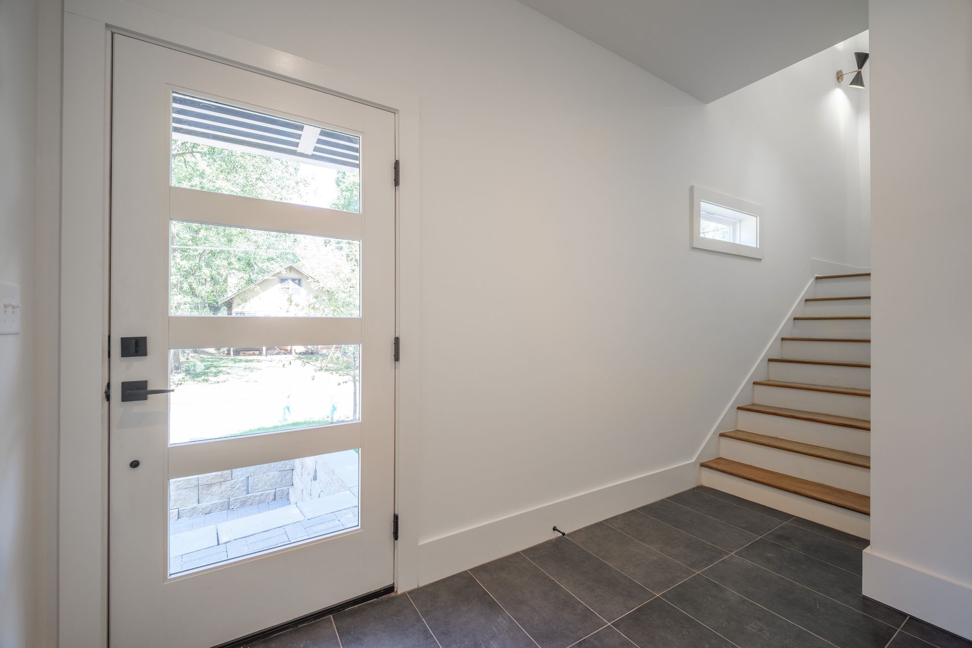 Entryway with a white door, stairs, and dark tile flooring. Sunlight streams through the glass door panels.