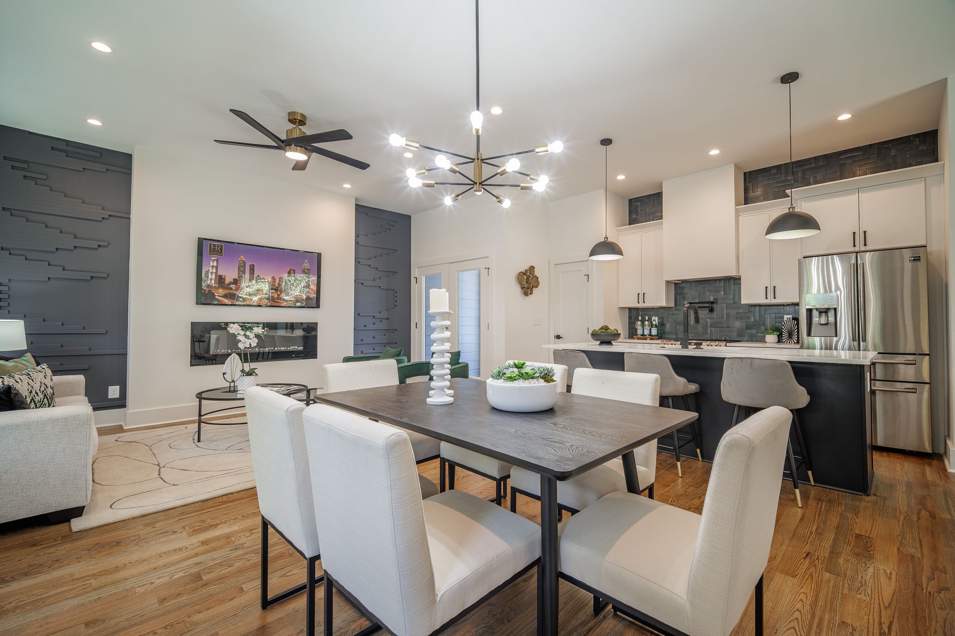 Open-concept living space with dining table, kitchen island, and fireplace. White, black, and wood tones throughout.