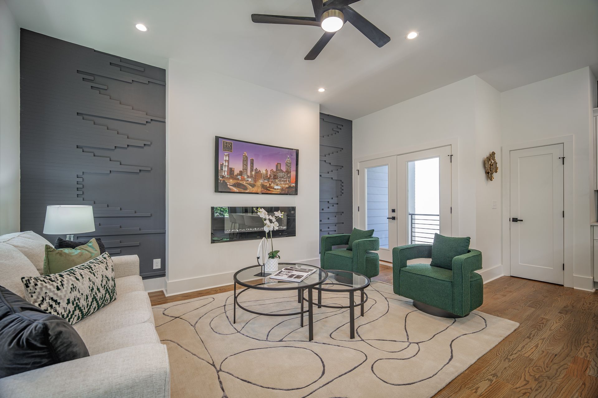 Living room with modern design: white sofa, green chairs, patterned rug, black accent wall, TV, and glass coffee table.