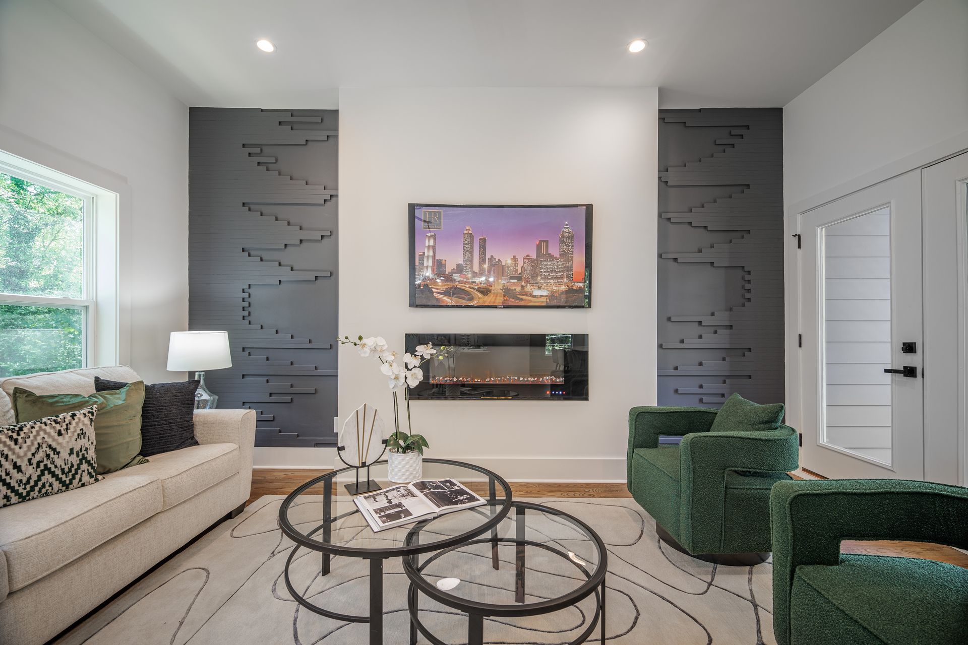 Living room with fireplace and mounted TV. Features a white sofa, green chairs, and glass coffee tables.