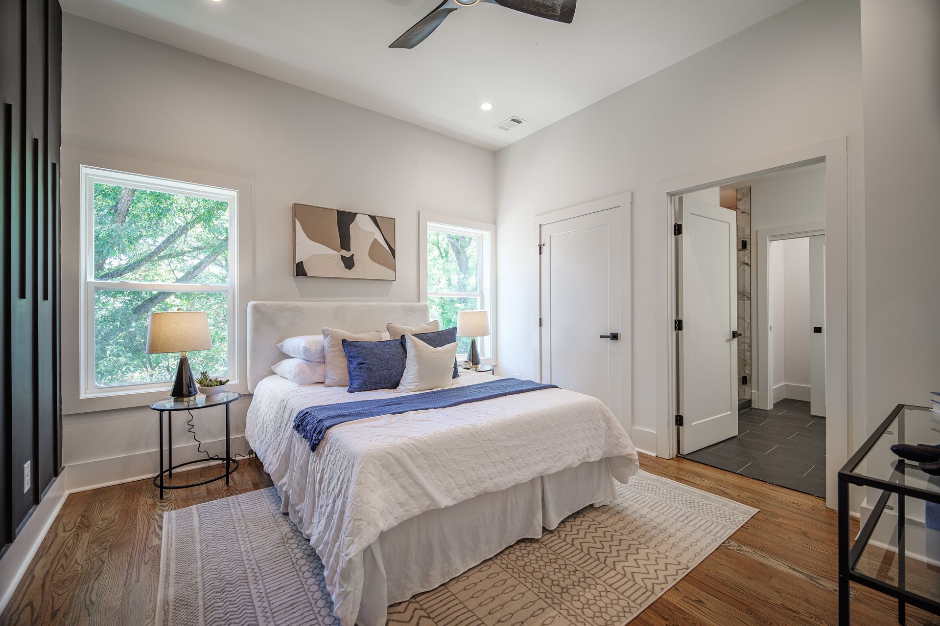 Bedroom with bed, windows, and two doors. White walls, wooden floor, neutral décor.