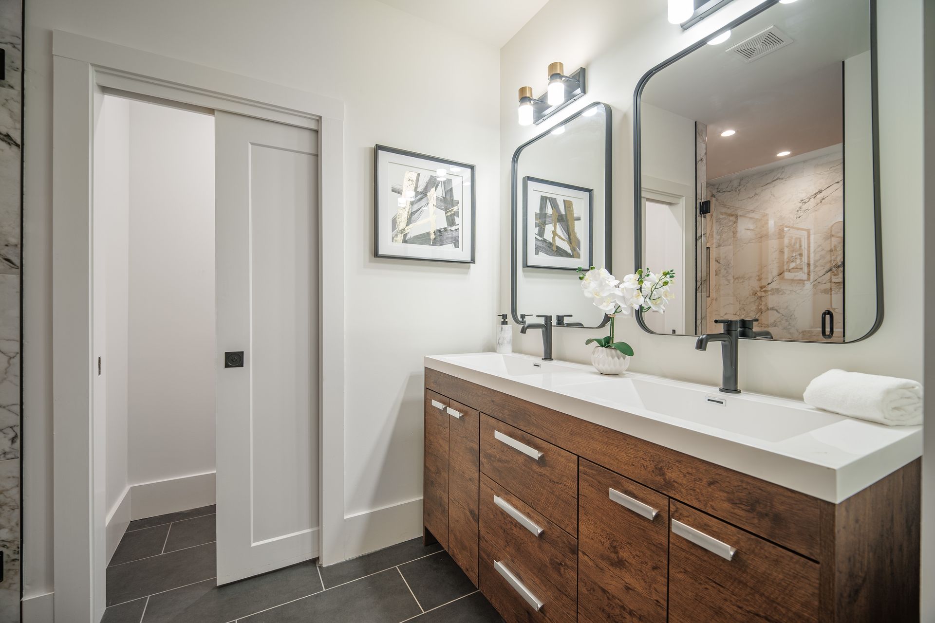 Modern bathroom with double vanity, two black-framed mirrors, and a wooden cabinet. Ajar door on left.