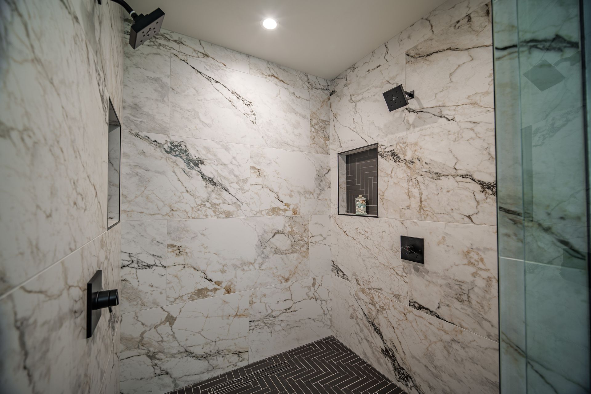 Shower with white marble walls, dark fixtures, and a dark tile floor. Glass shower door is on the right.