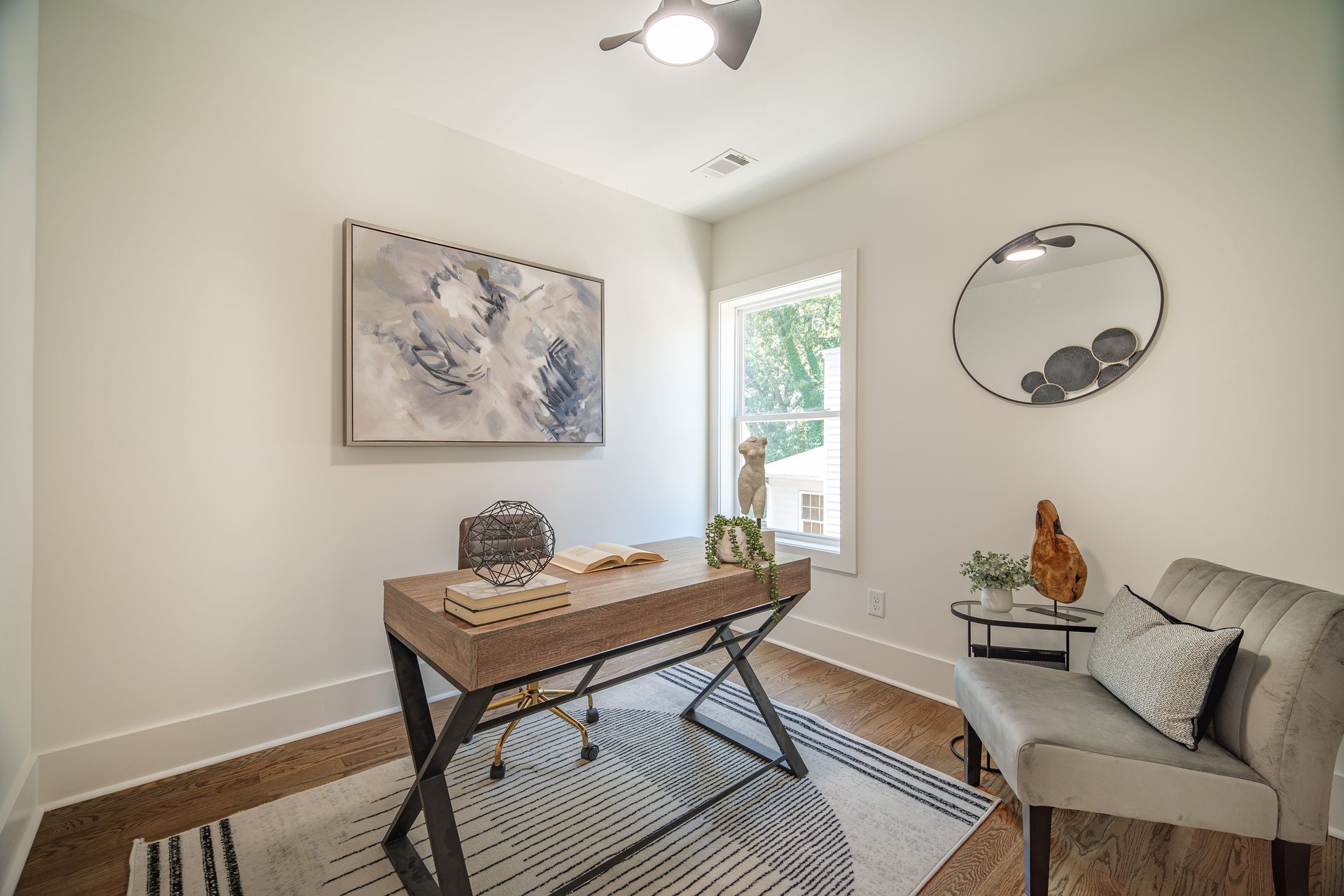 Home office with desk, chair, artwork, rug, mirror, and a small side table near a window.