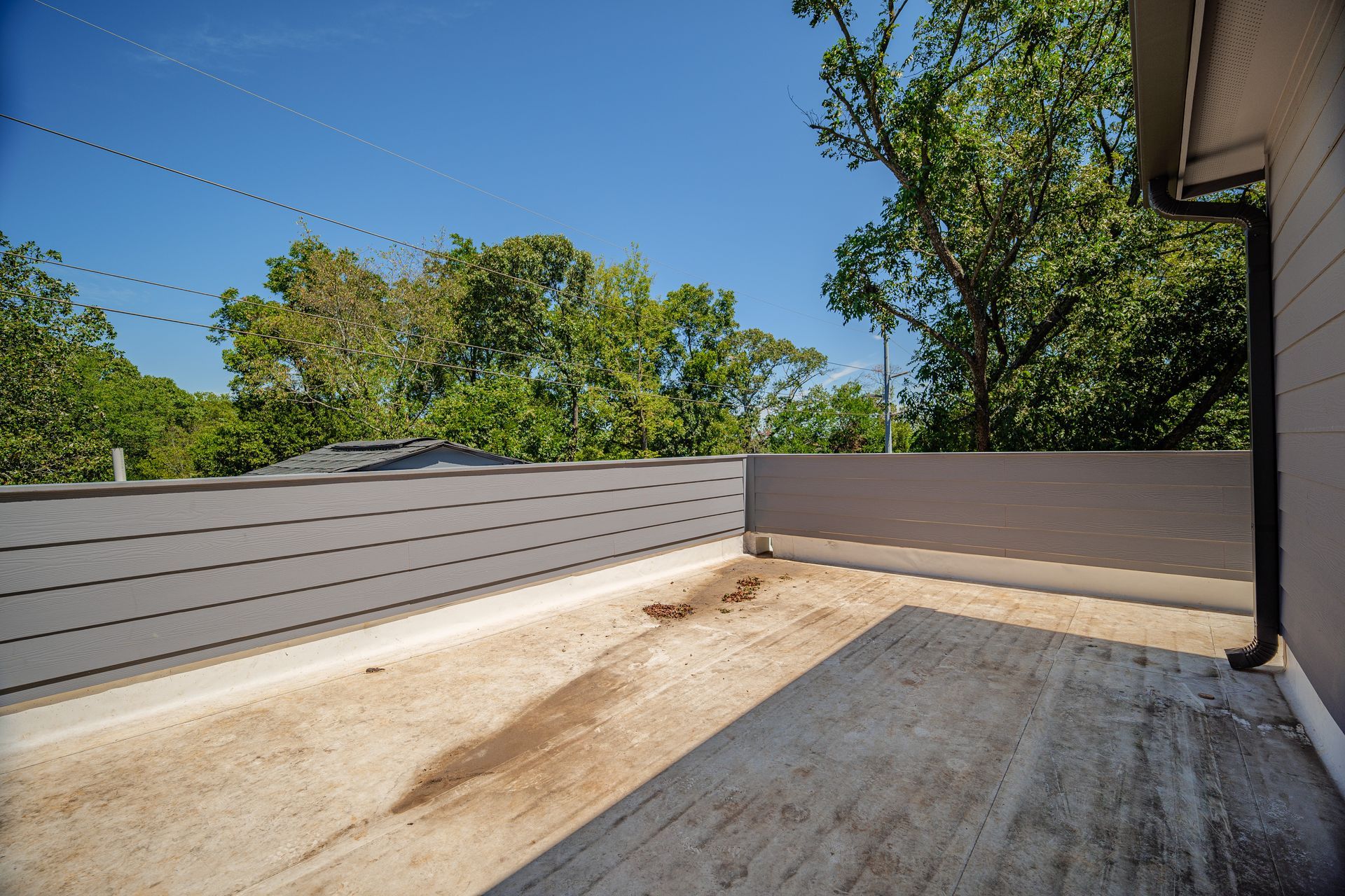 Rooftop patio with gray siding, light-colored flat surface, brown stain, trees, and a clear blue sky.