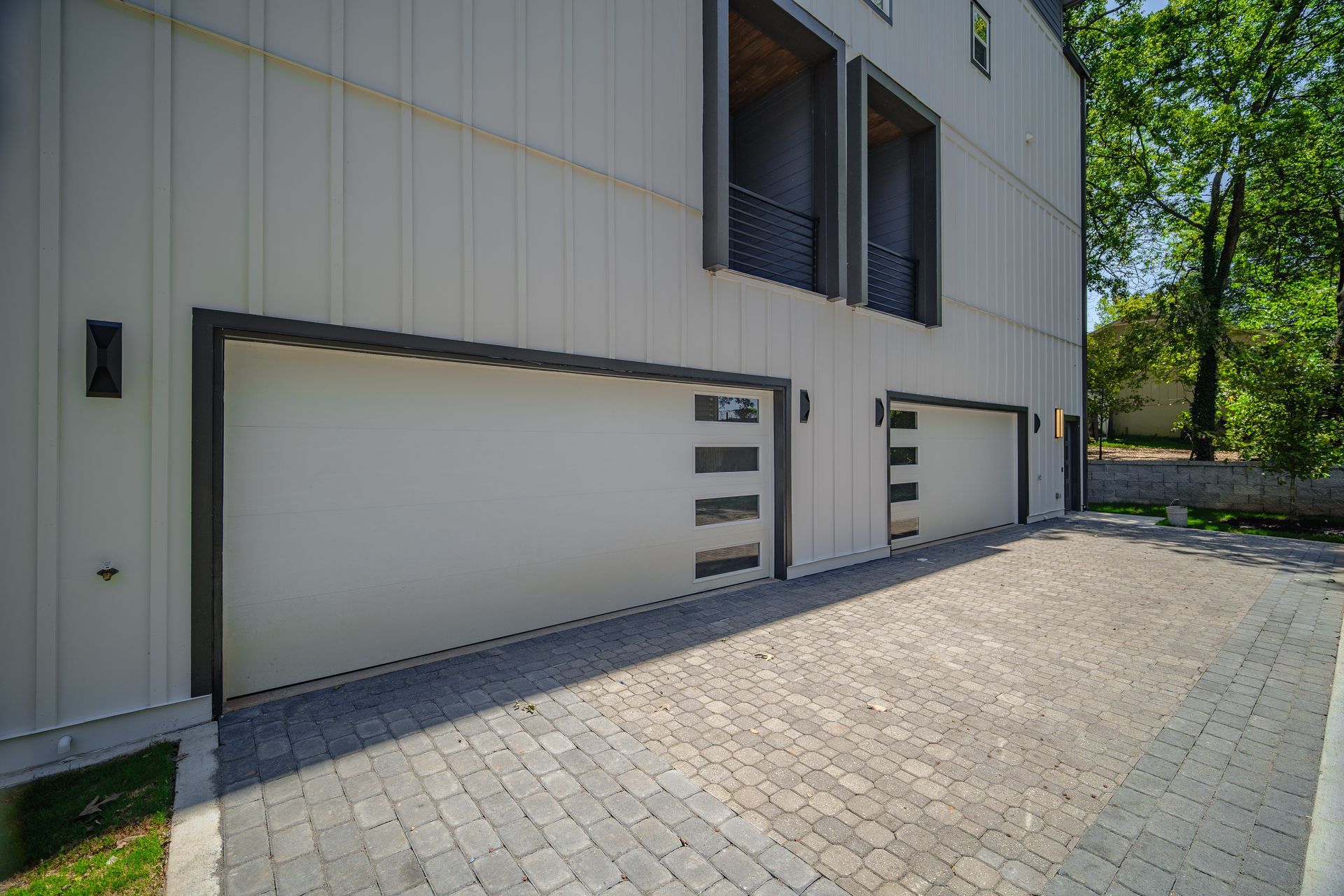 Two white garage doors with black trim, on a light-colored building. Paved driveway, trees in background.