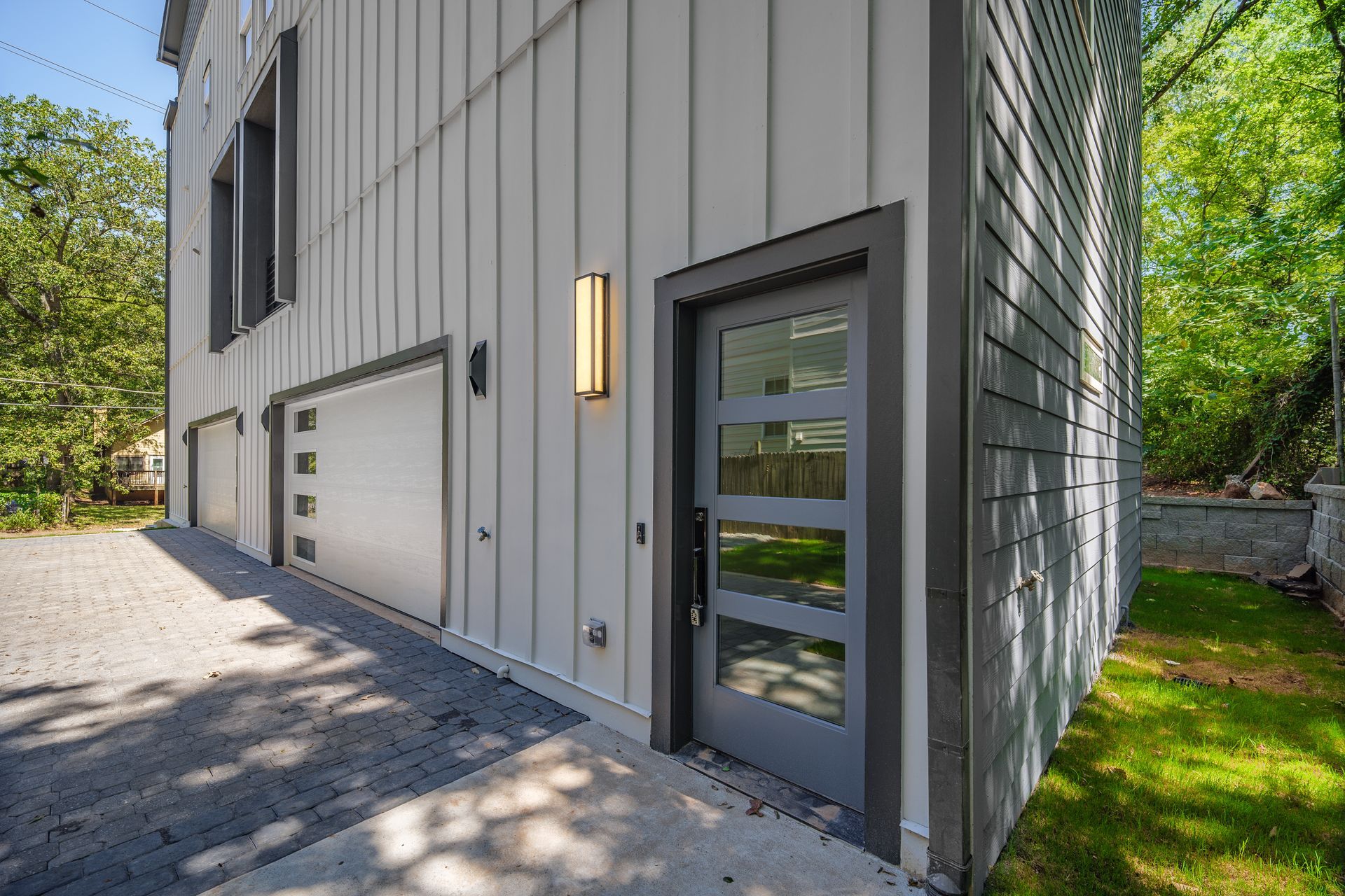 Modern gray house exterior with garage doors, entrance door, and a brick driveway.