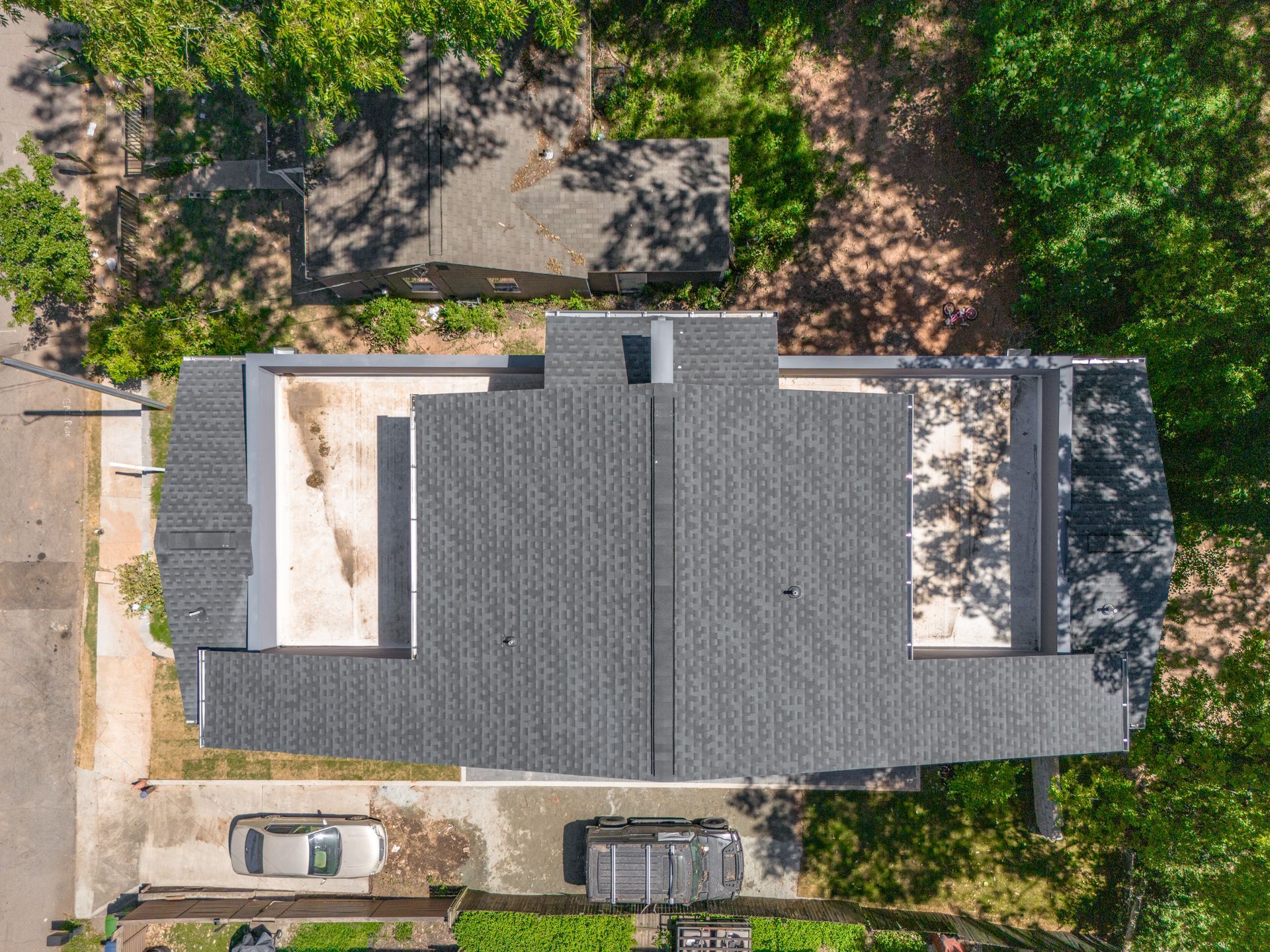 Overhead view of a modern house with a dark gray shingled roof, light gray flat roof sections, and surrounding greenery.