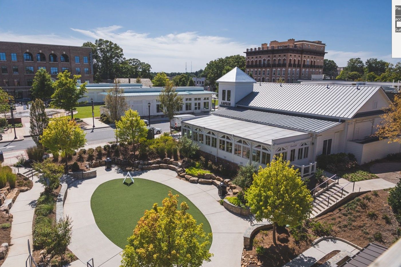 Aerial view of a white building with a courtyard, green lawn, trees, and other buildings on a sunny day.