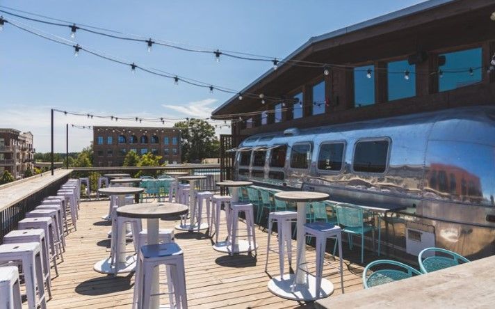 Rooftop bar with white tables and stools. Shiny airstream trailer building. Sunny day.
