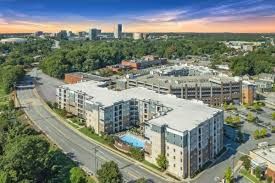 Aerial view of apartment complex with pool, trees, road, and city skyline in the background.