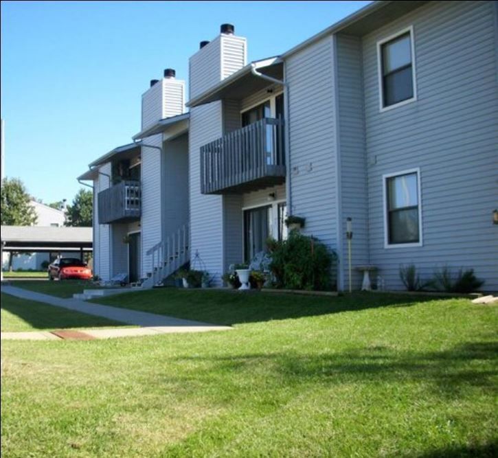 Apartment building with light blue siding, balconies, green lawn, and a car in the background.