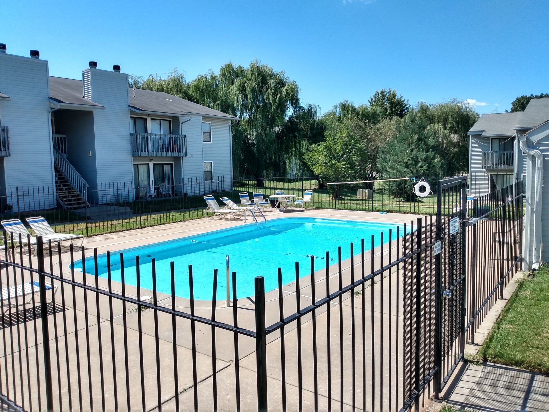 Swimming pool area with apartment buildings, sunny day. Fence surrounds blue water. Lounge chairs visible.