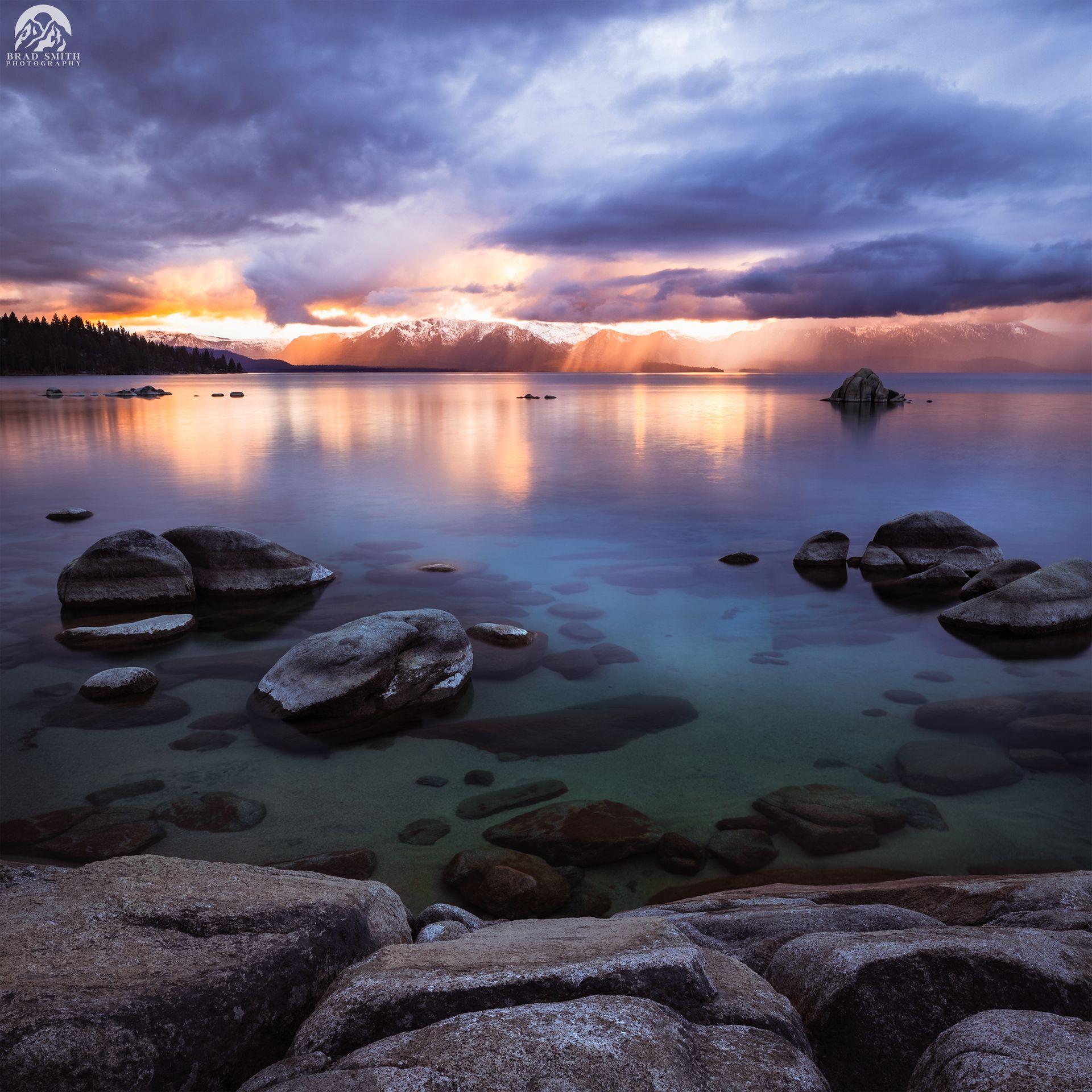 Rocky shoreline at sunset with calm reflective water, dramatic purple clouds, and warm light on the horizon
