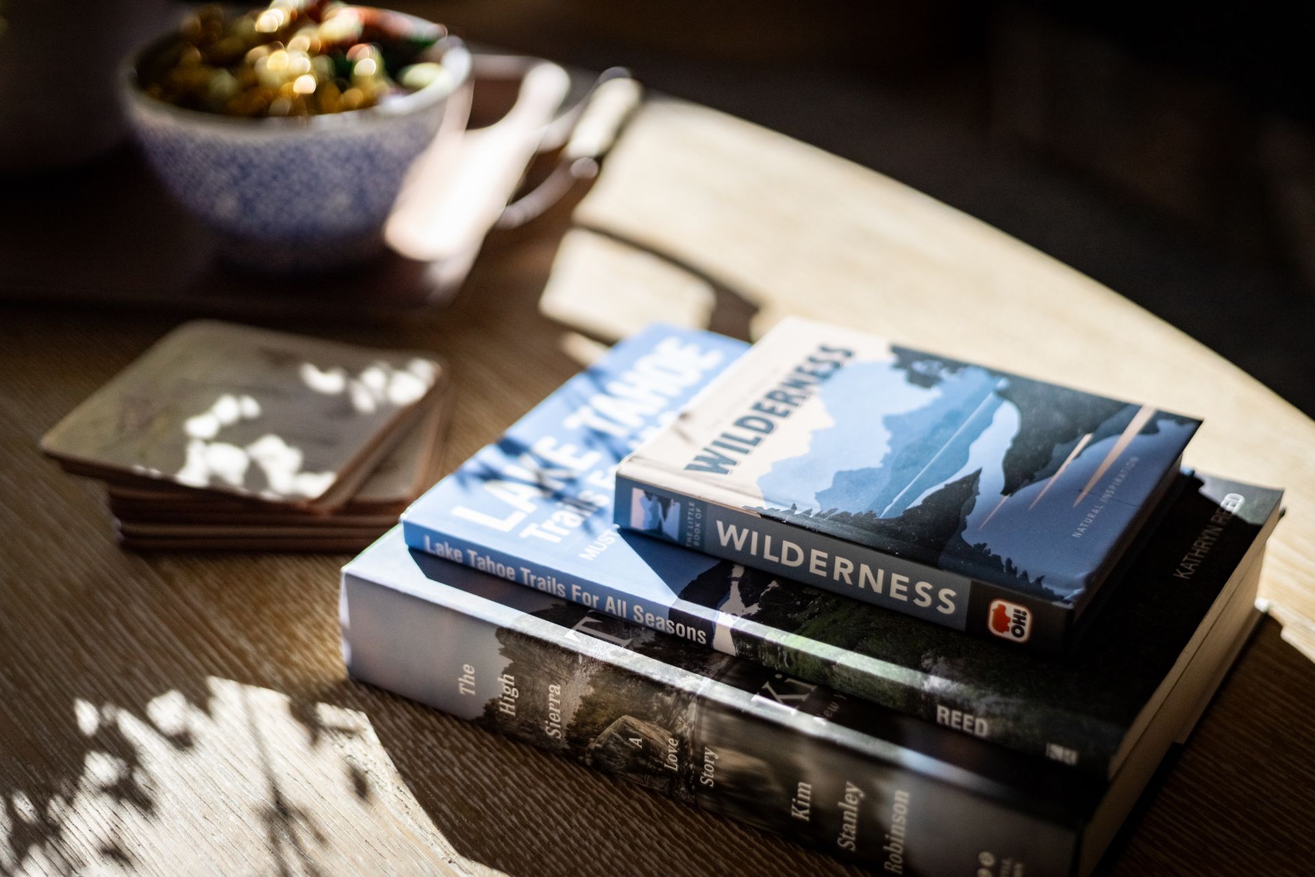 Stack of books on a wooden table beside a bowl of fruit in warm sunlight.