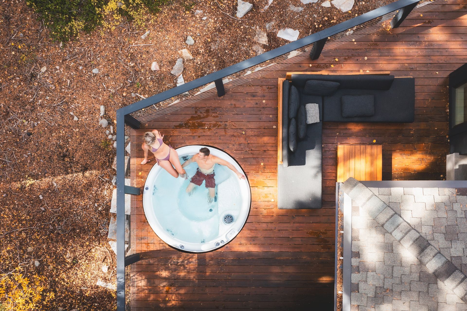 Person relaxing in a round hot tub on a wooden deck beside an outdoor sectional sofa.