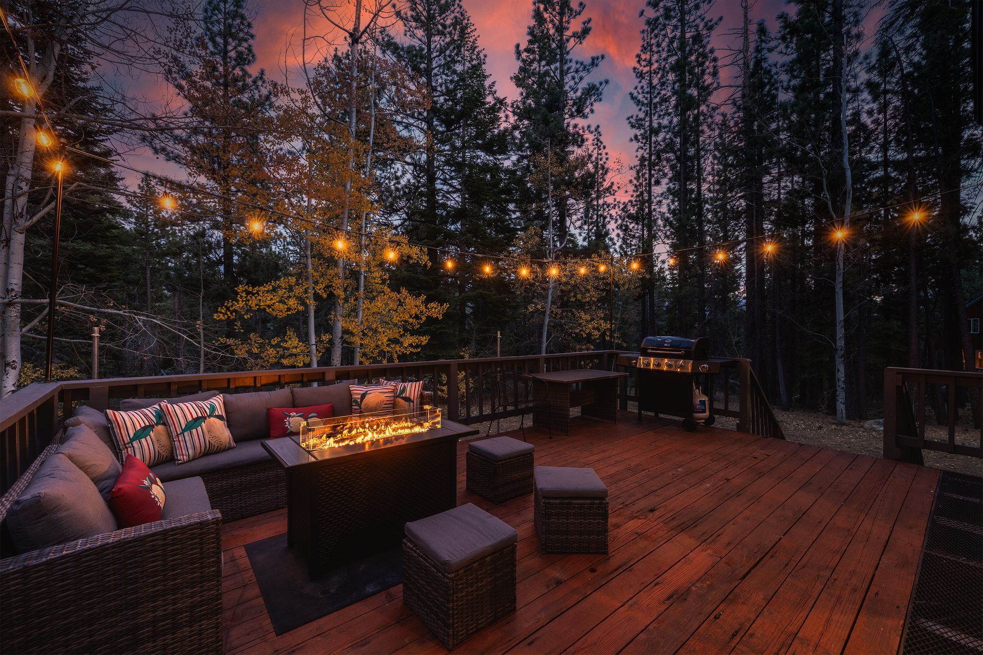 Cozy outdoor deck at dusk with fire pit, sectional seating, and string lights overlooking a forest