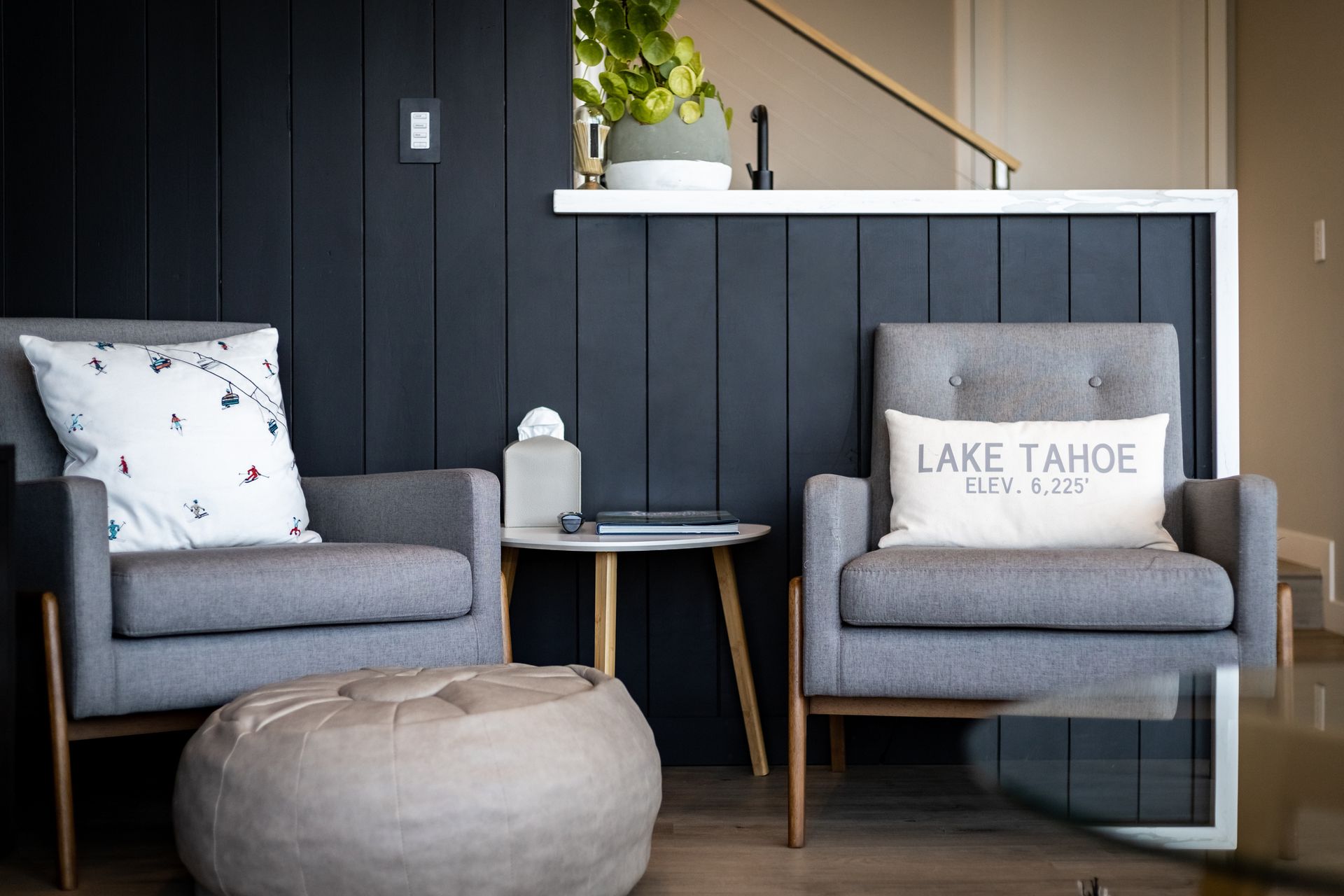 Modern lounge with two gray chairs, a round ottoman, and a white Lake Tahoe pillow against a dark wall