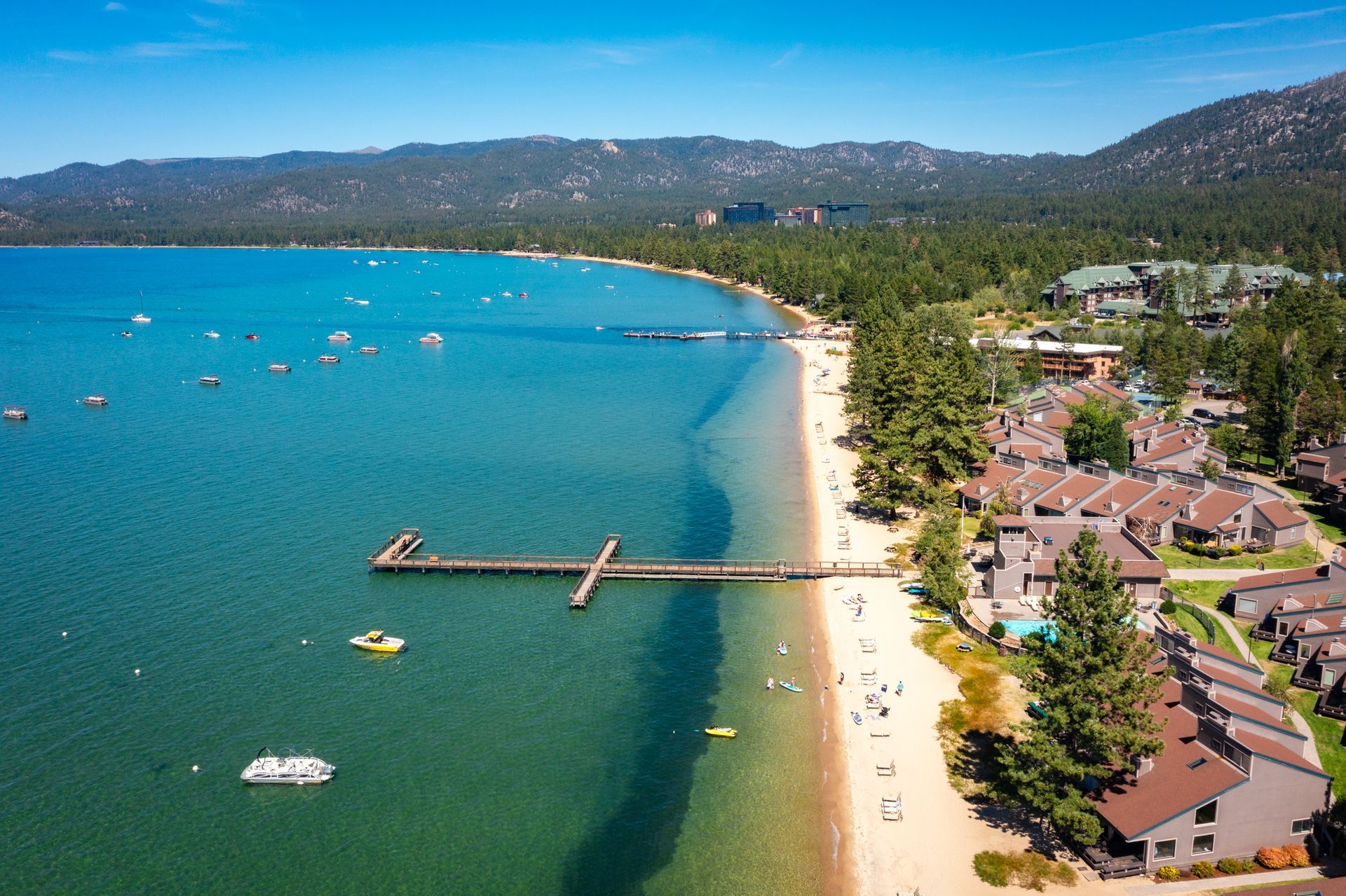 Aerial view of a turquoise lake, sandy beach, pier, and lakeside cottages with mountains in the distance