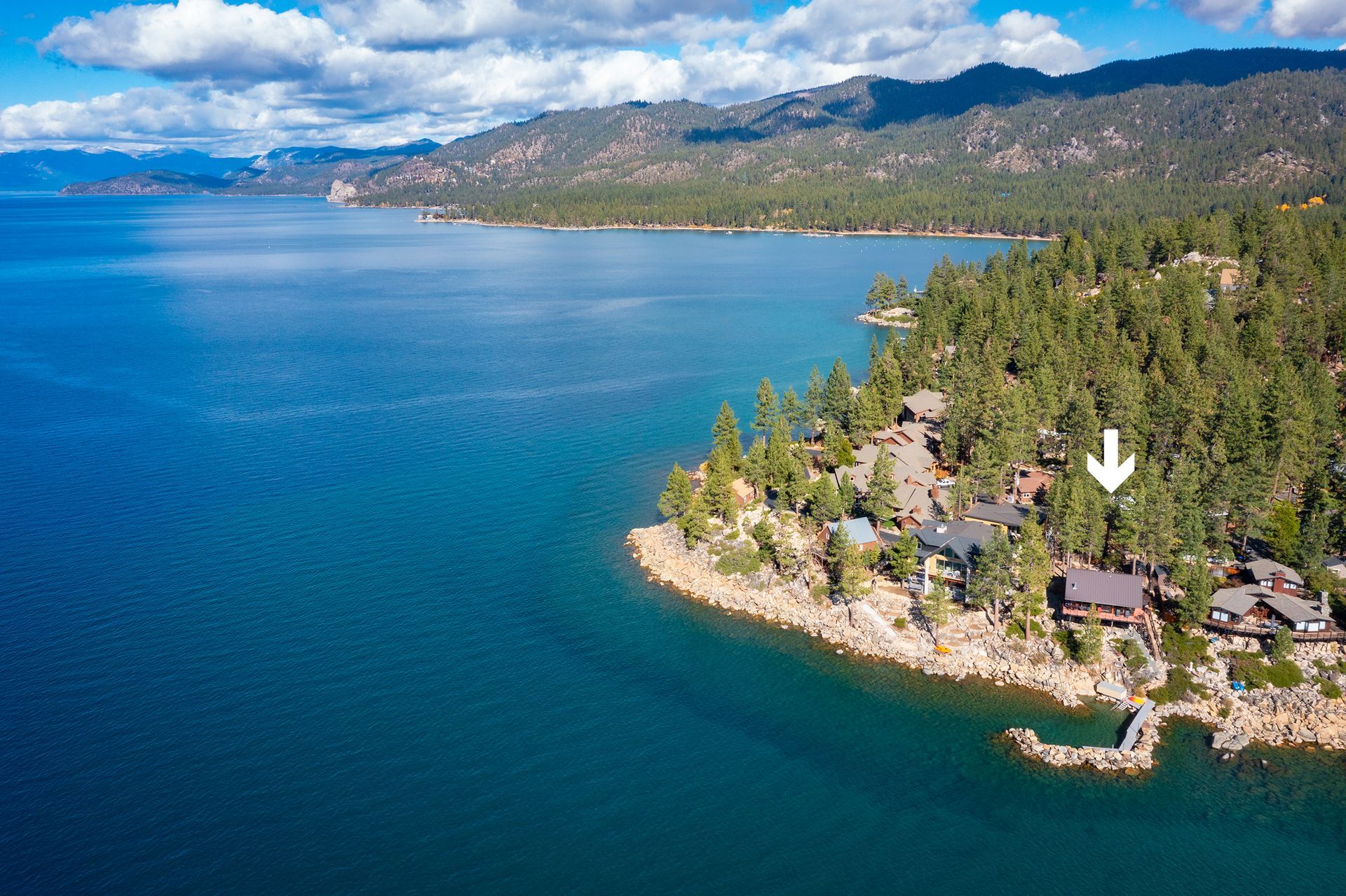Aerial view of a blue lake beside a forested shoreline with houses and mountains in the distance