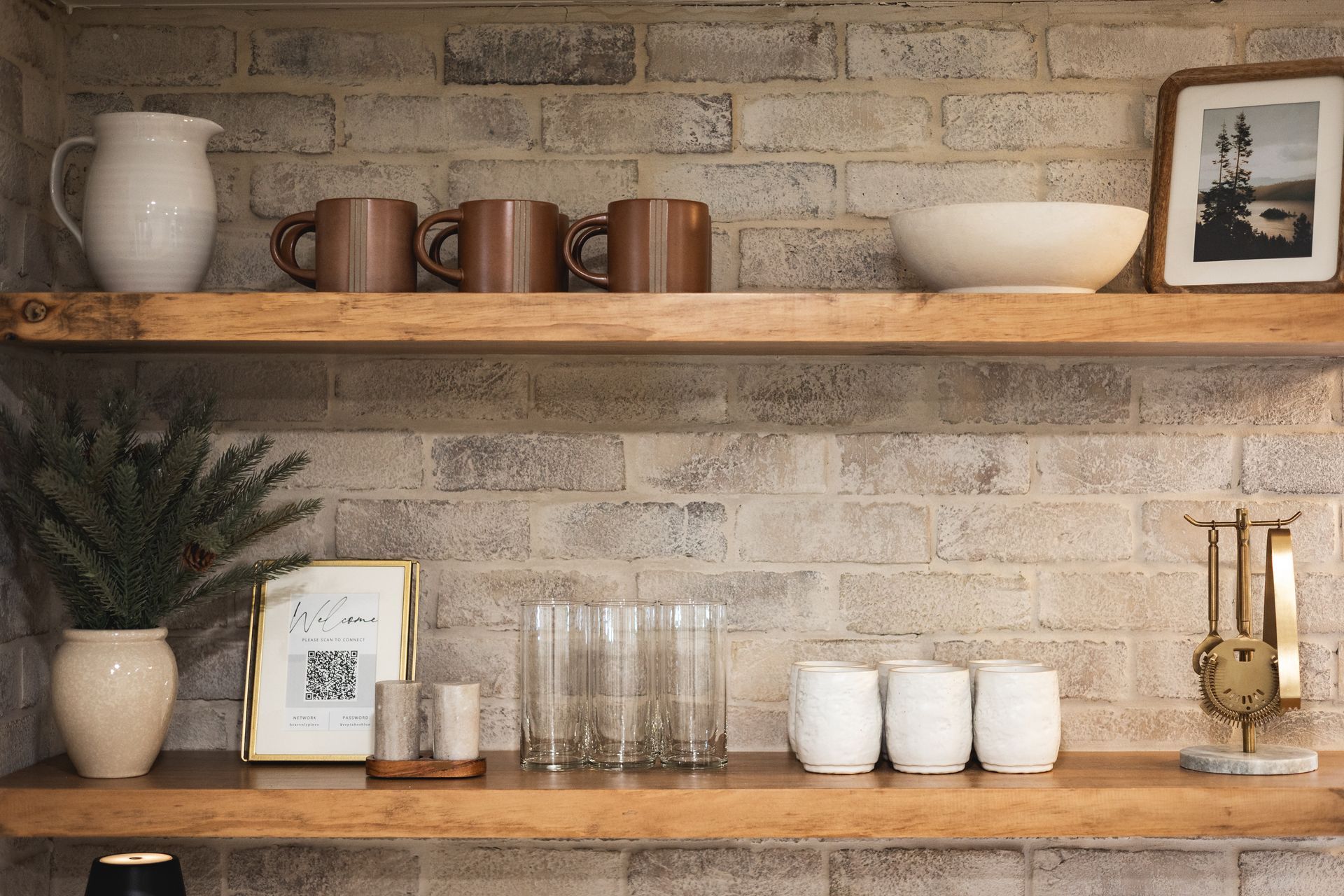 Rustic shelves with ceramic mugs, glasses, bowls, a plant, and framed decor against a brick wall