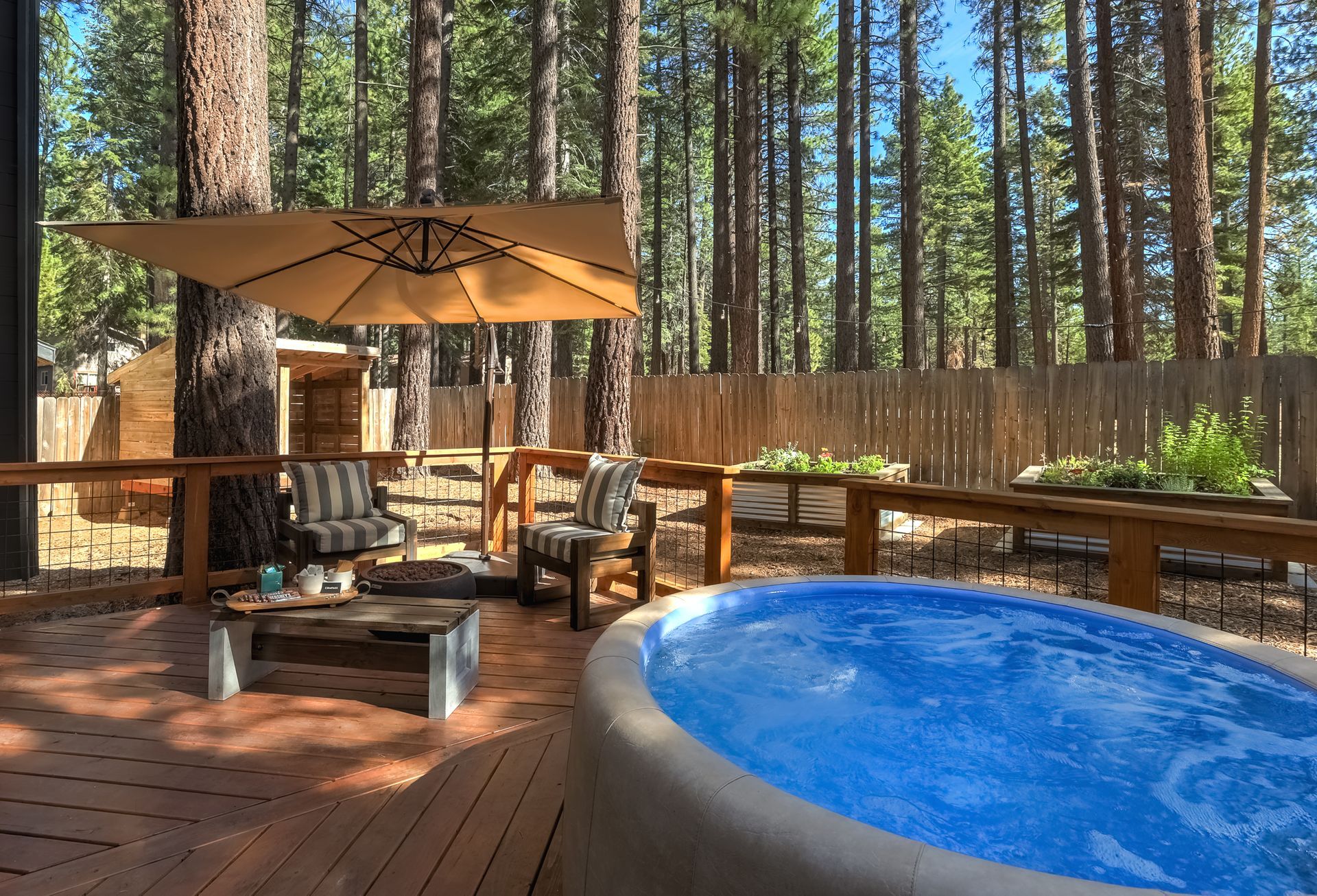 Wooden deck with hot tub, patio seating, and tan umbrella in a pine forest backyard